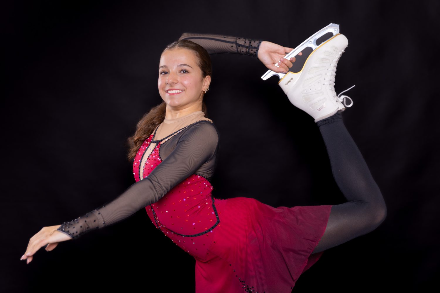 Eine Sportlerin in einem rot-weißen Sportoberteil und schwarzen Shorts kniet in einer Sprint-Startposition und lächelt vor einem schlichten schwarzen Hintergrund in die Kamera - und verdeutlicht so den Schub, den Sie mit Aronia Shot spüren.