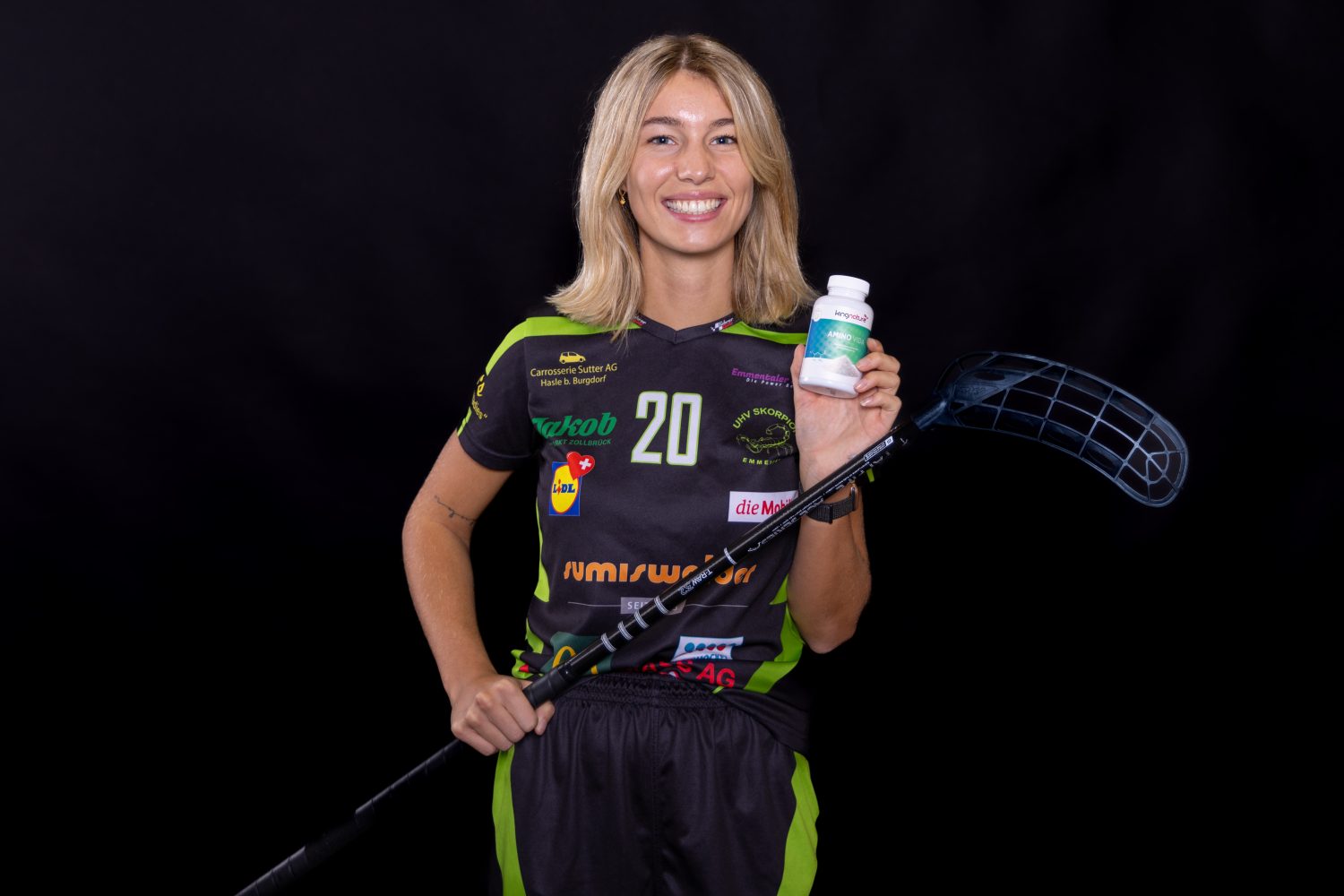 Eine lächelnde Frau in einem schwarz-grünen Sportdress hält einen Unihockeyschläger und zeigt vor einem schwarzen Hintergrund eine Flasche mit Amino Vida-Ergänzungsmitteln.