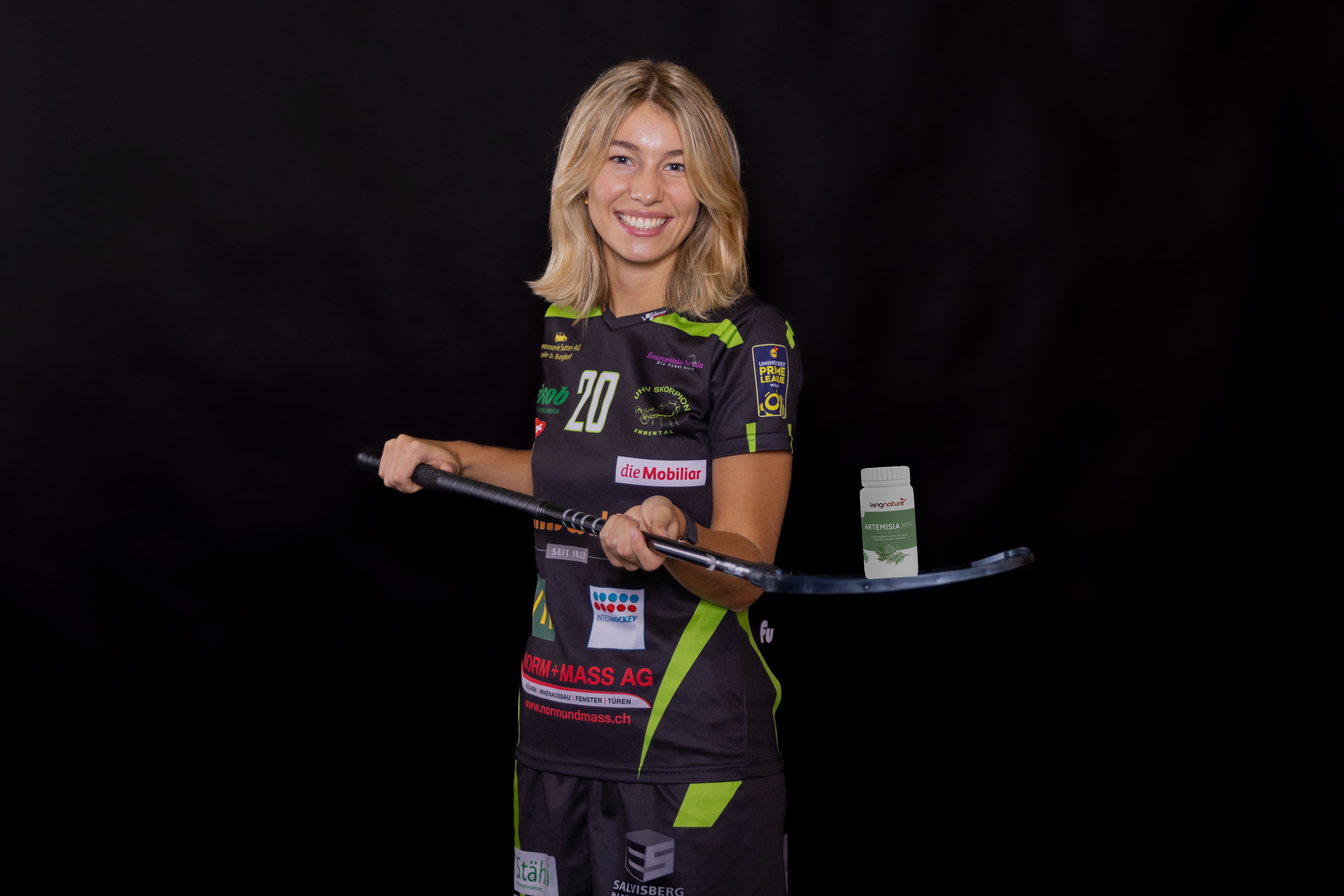 Eine lächelnde Frau in einem schwarz-grünen Artemisia-Mini-Bio-Sportdress hält einen Unihockeyschläger waagerecht vor sich auf einem schlichten schwarzen Hintergrund.