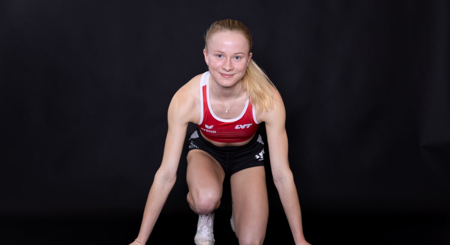 Eine Sportlerin in einem rot-weißen Sportoberteil und schwarzen Shorts kniet in einer Sprint-Startposition und lächelt vor einem schlichten schwarzen Hintergrund in die Kamera - und verdeutlicht so den Schub, den Sie mit Aronia Shot spüren.