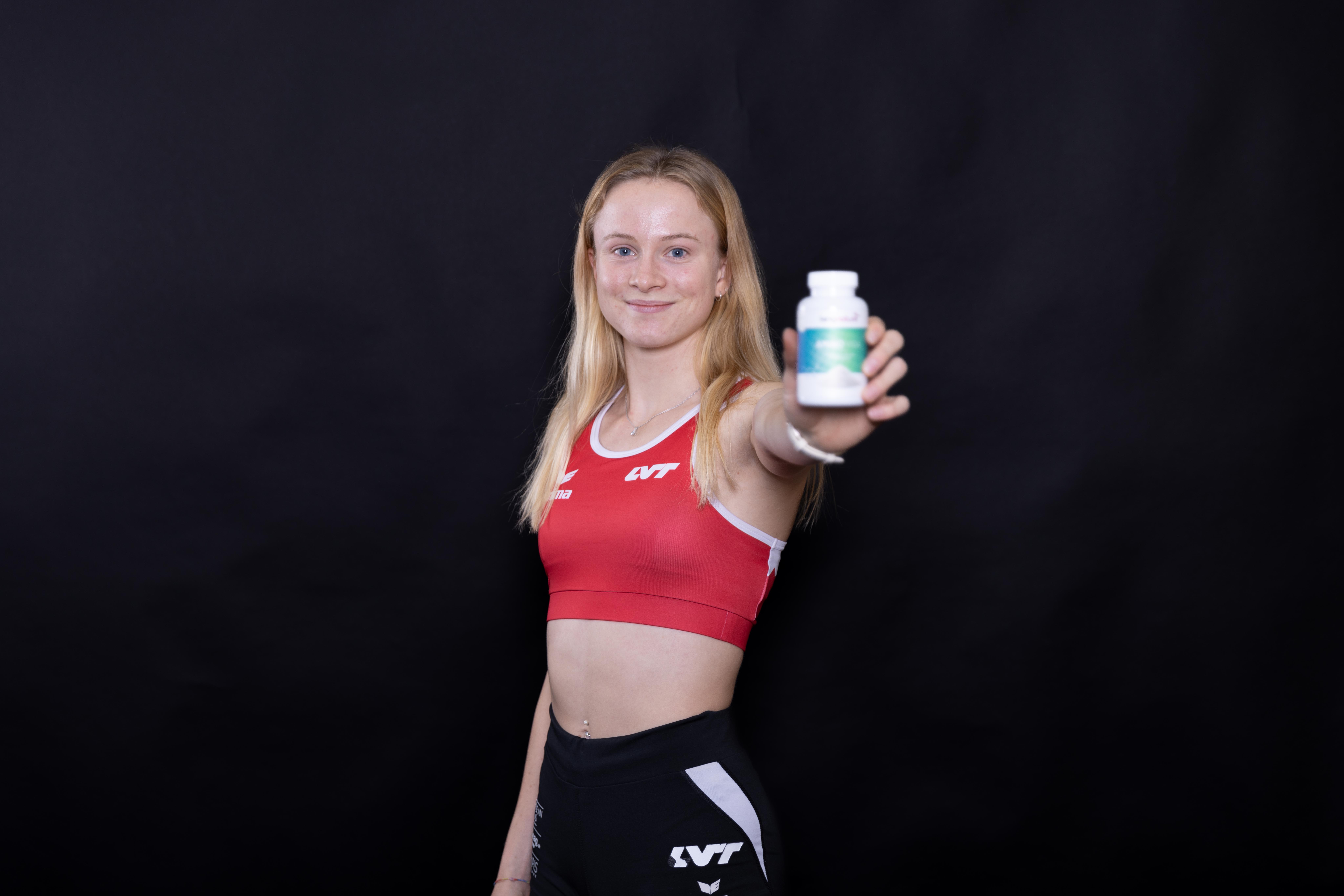 Linda Bichsel steht in sportlicher Kleidung vor einem schwarzen Hintergrund, lächelt und hält eine weiße Flasche mit dem Etikett in Richtung Kamera.