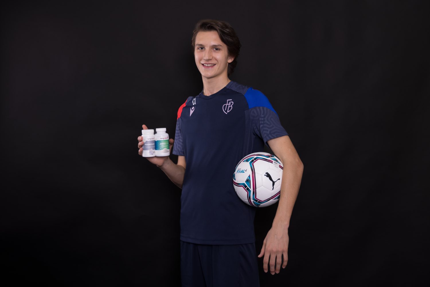 Ein junger Mann in einem marineblauen Sportdress hält einen Fußball unter einem Arm und drei Amino Vida-Flaschen in der anderen Hand. Er steht vor einem schlichten schwarzen Hintergrund und lächelt in die Kamera.