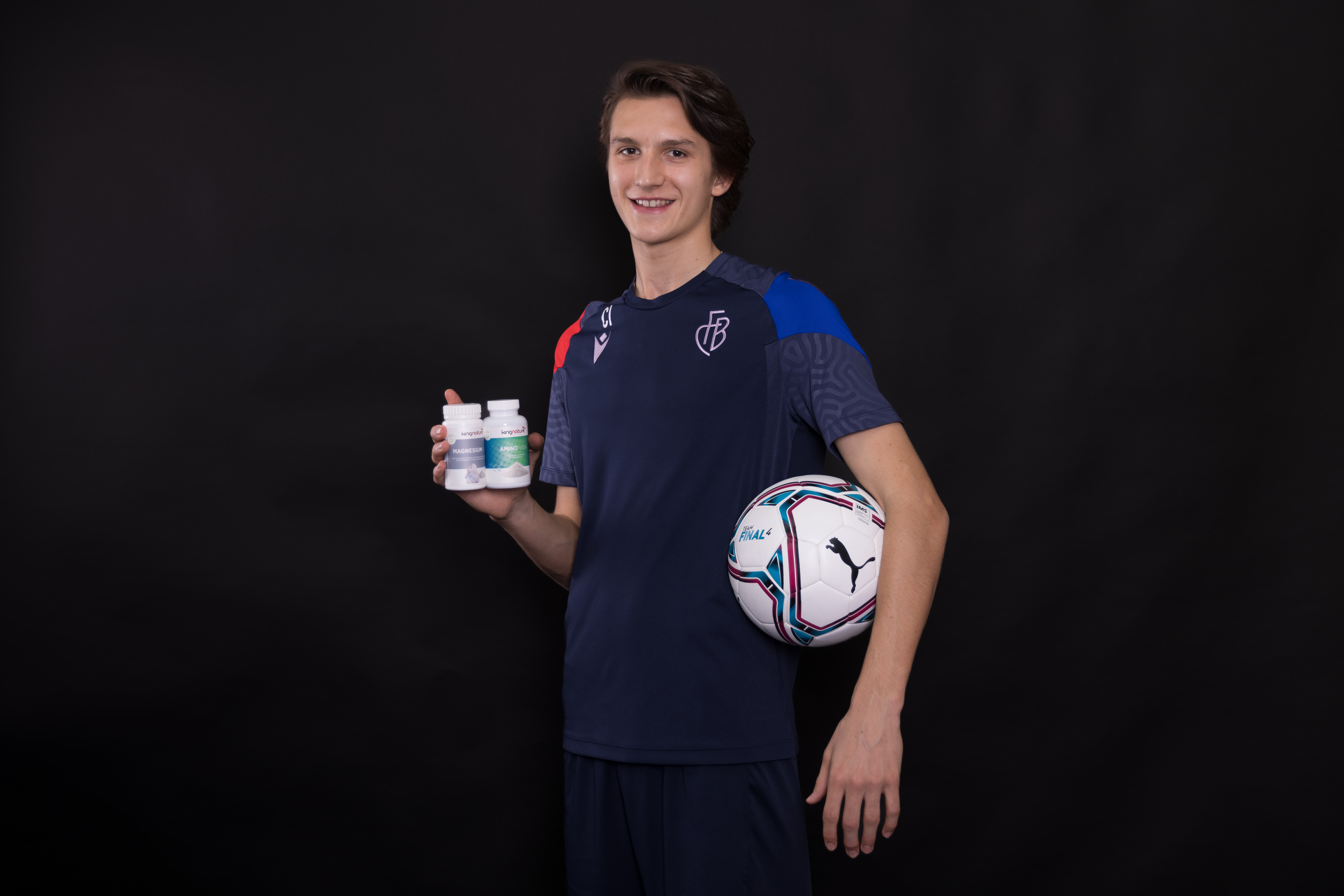 Ein junger Mann in einem marineblauen Sportdress hält einen Fußball unter einem Arm und drei Amino Vida-Flaschen in der anderen Hand. Er steht vor einem schlichten schwarzen Hintergrund und lächelt in die Kamera.