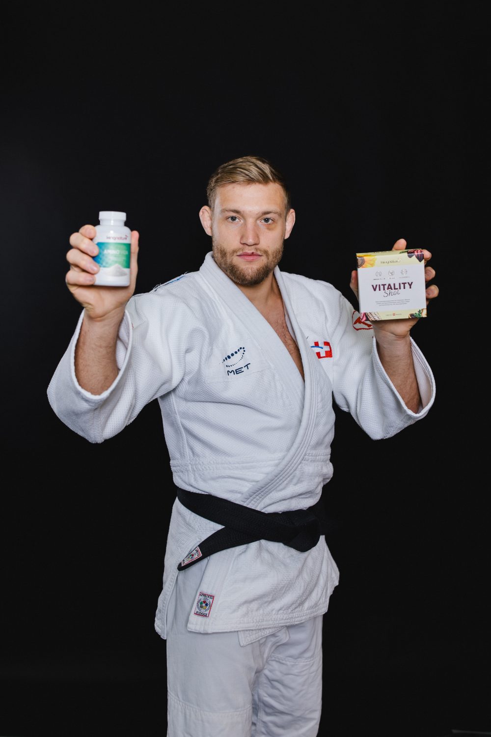 Ein Mann in weißem Judoanzug und schwarzem Gürtel steht vor einem schwarzen Hintergrund und hält eine Flasche Amino Vida in der einen und eine Schachtel mit der Aufschrift Vitality in der anderen Hand, beide in Richtung der Kamera erhoben.