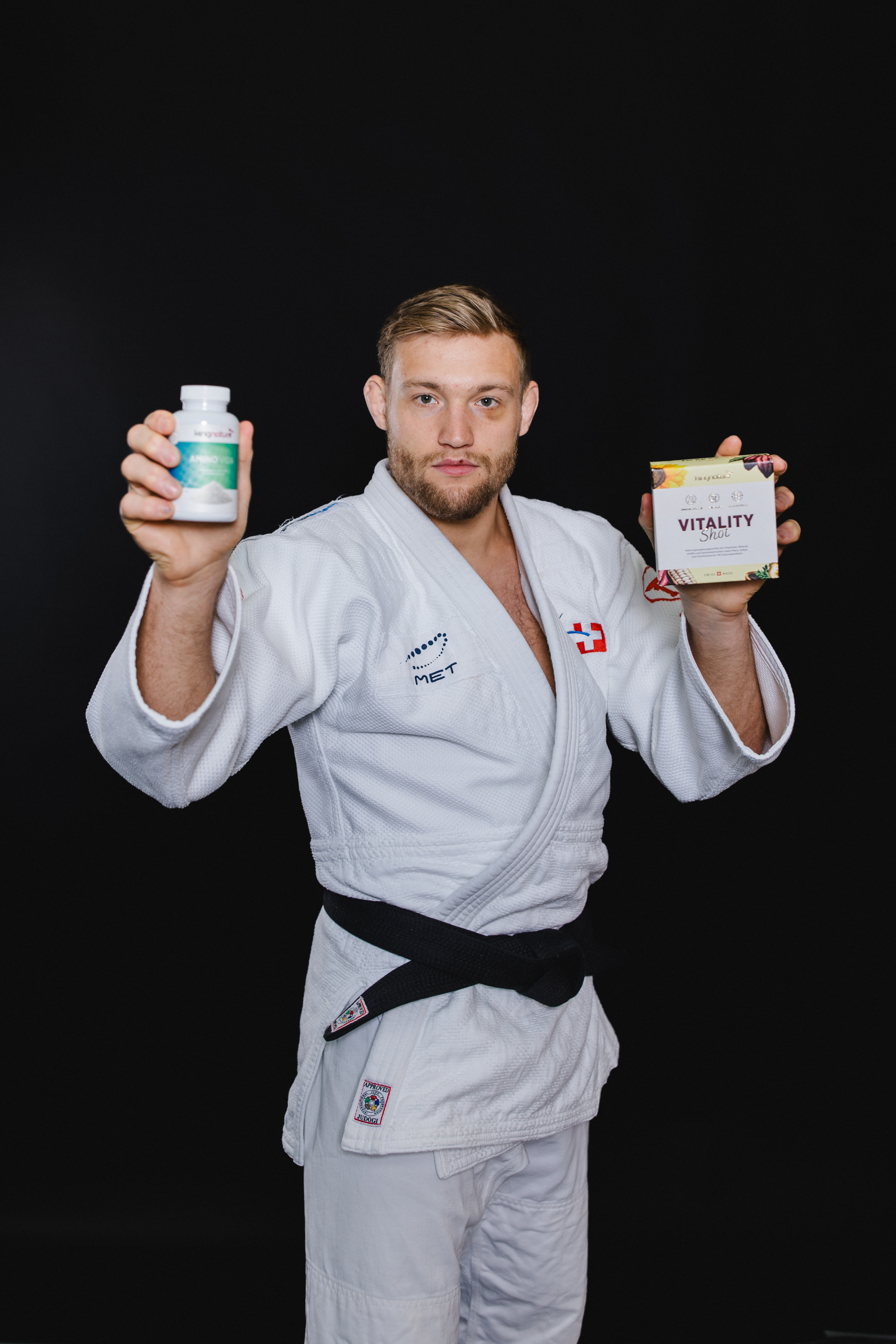Ein Mann in weißem Judoanzug und schwarzem Gürtel steht vor einem schwarzen Hintergrund und hält eine Flasche Amino Vida in der einen und eine Schachtel mit der Aufschrift Vitality in der anderen Hand, beide in Richtung der Kamera erhoben.