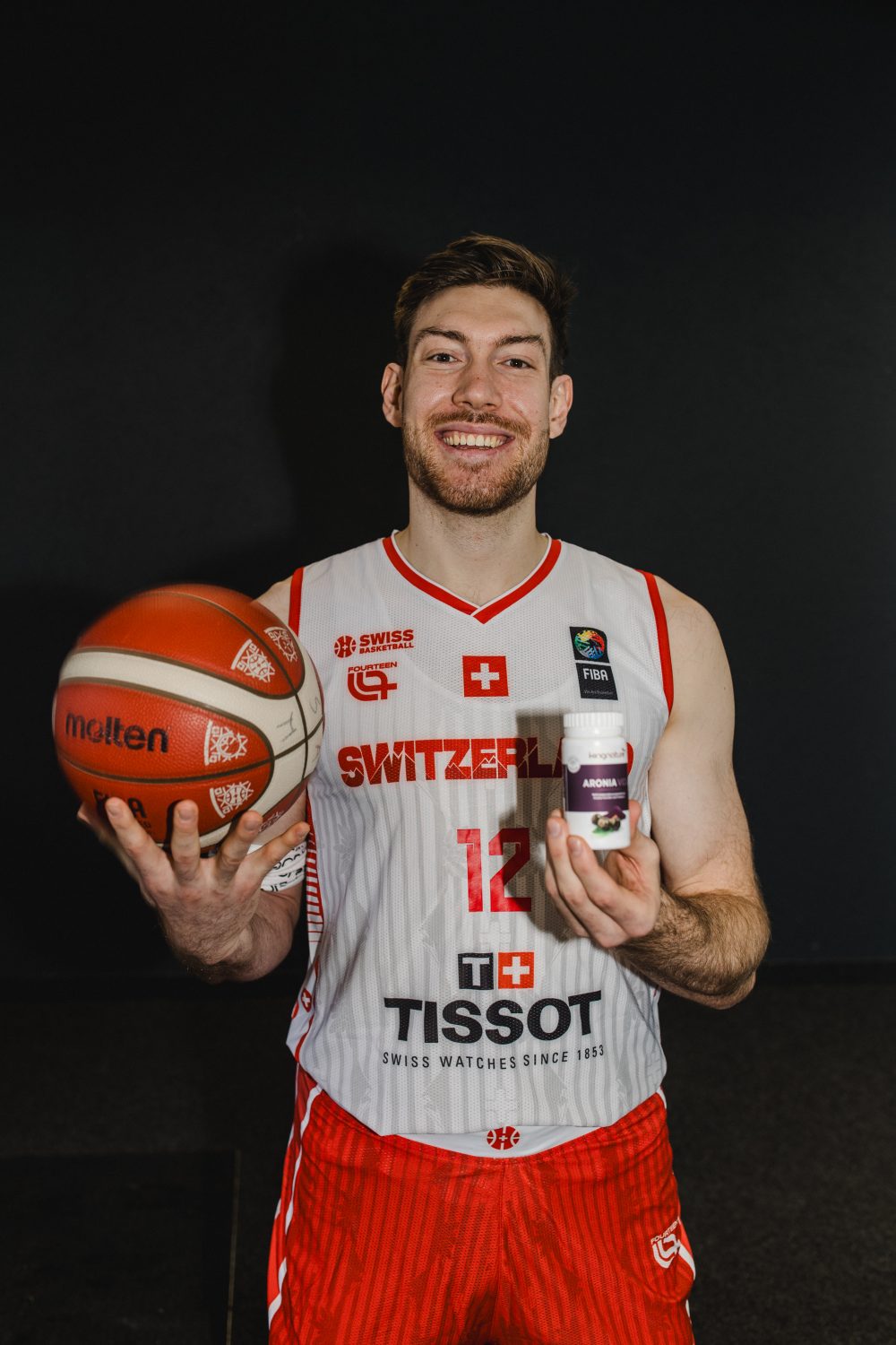 Ein lächelnder Basketballspieler im Schweizer Trikot hält vor einem dunklen Hintergrund in der einen Hand einen Basketball und in der anderen eine Flasche mit Aronia Vida-Nahrungsergänzungsmitteln.