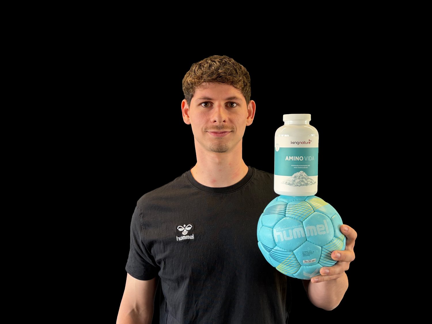 Vor einem schwarzen Hintergrund hält ein Mann in einem schwarzen Hummel-Shirt einen blauen Hummel-Fußball in der Hand, auf dem eine Flasche Amino Vida-Nahrungsergänzungsmittel balanciert – ein Bild, das Levin Wanners Leidenschaft für Fitness und Sport zum Ausdruck bringt.