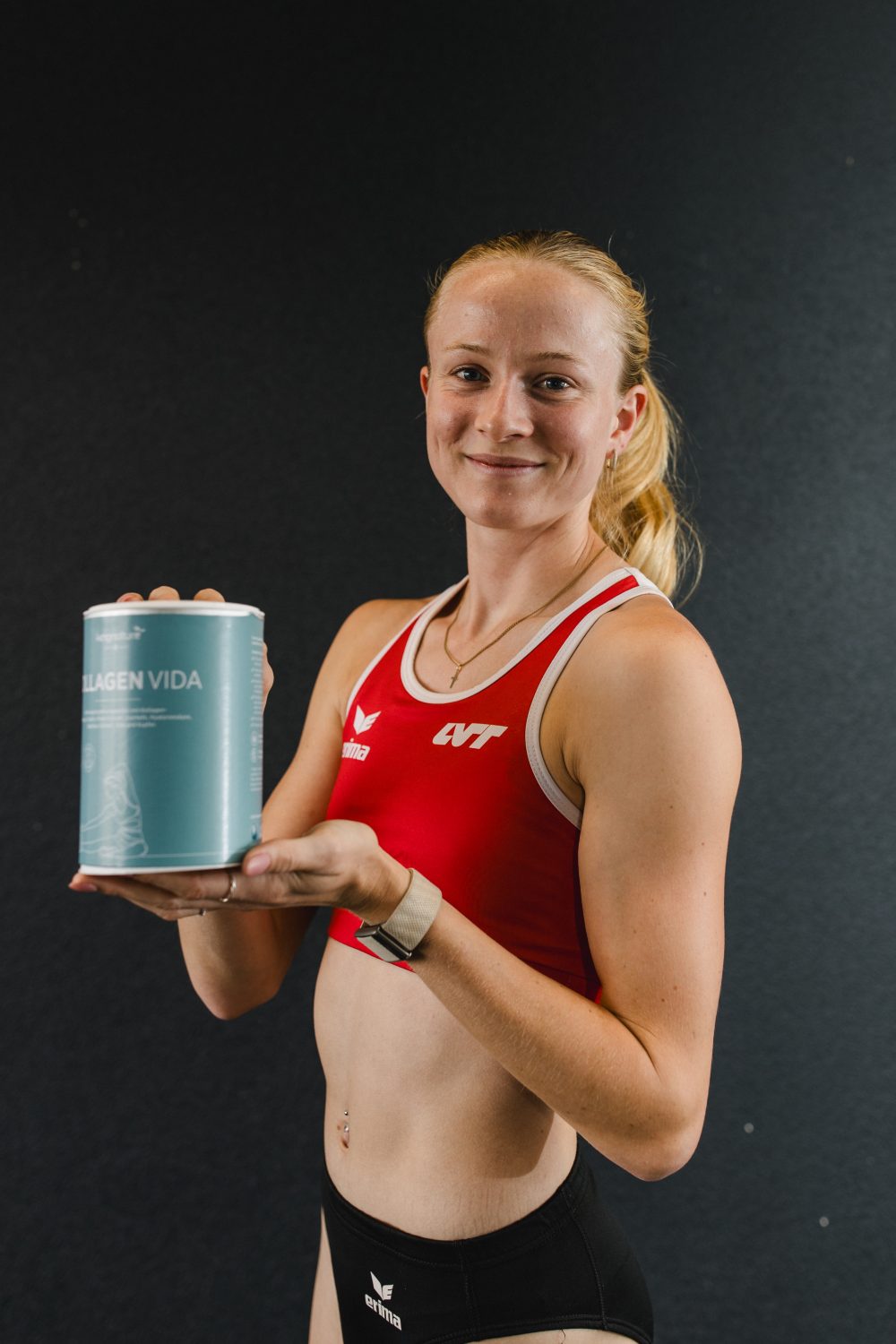 Eine lächelnde Frau in einem roten Sport-BH hält eine Packung „Collagen Vida“ in der Hand und hebt vor einem dunklen Hintergrund deren Vorteile hervor.