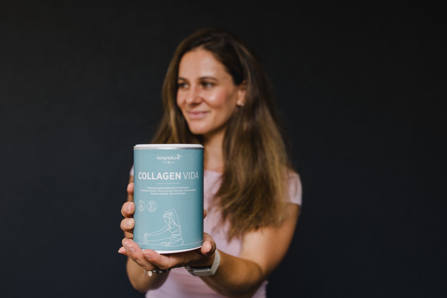 Eine Frau in einem hellen Shirt hält einen blauen Behälter mit „Collagen Vida“ in Richtung Kamera und lächelt sanft. Auf dem Produktetikett sind Text und eine Strichzeichnung vor einem schlichten dunklen Hintergrund zu sehen.