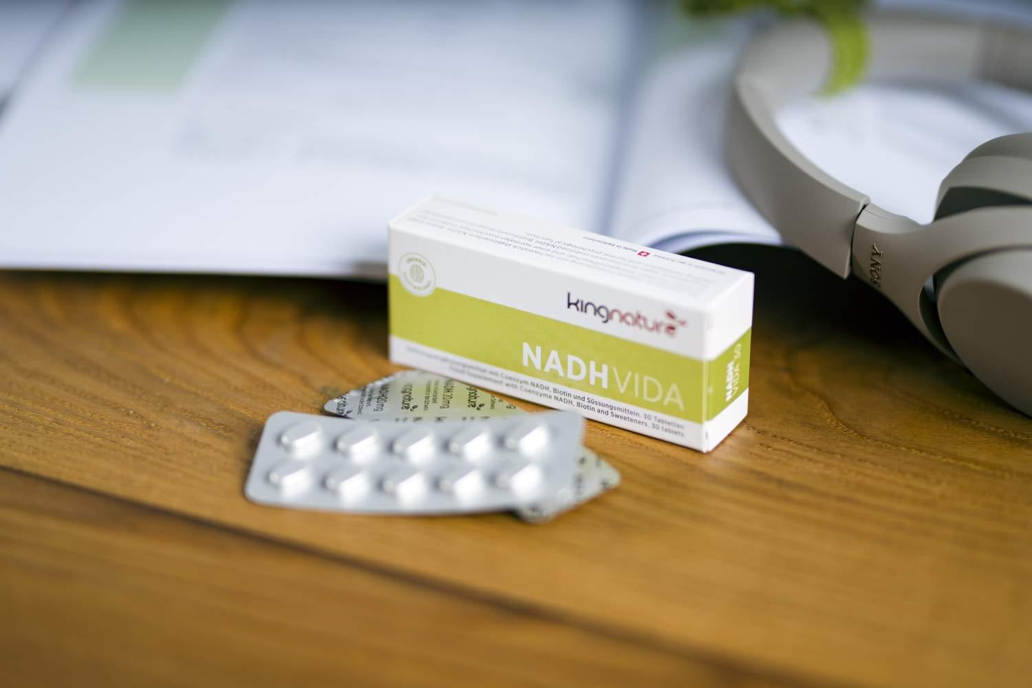 Eine Schachtel mit der Aufschrift „NADH VIDA“ und Blisterpackungen mit Tabletten stehen auf einem Holztisch. Im Hintergrund sind ein verschwommenes offenes Buch und Kopfhörer zu sehen, die Konzentration und PEP-Protokoll-Energie suggerieren.