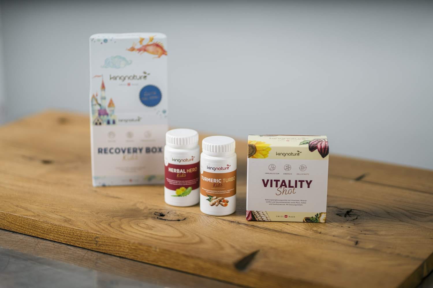 Drei Nahrungsergänzungsmittel von kingnature – Recovery Box Kids, Herbal Hero Kids und Vitality Shot – werden auf einer Holzoberfläche vor einem neutralen Hintergrund präsentiert, wodurch ihre natürlichen Inhaltsstoffe hervorgehoben werden.