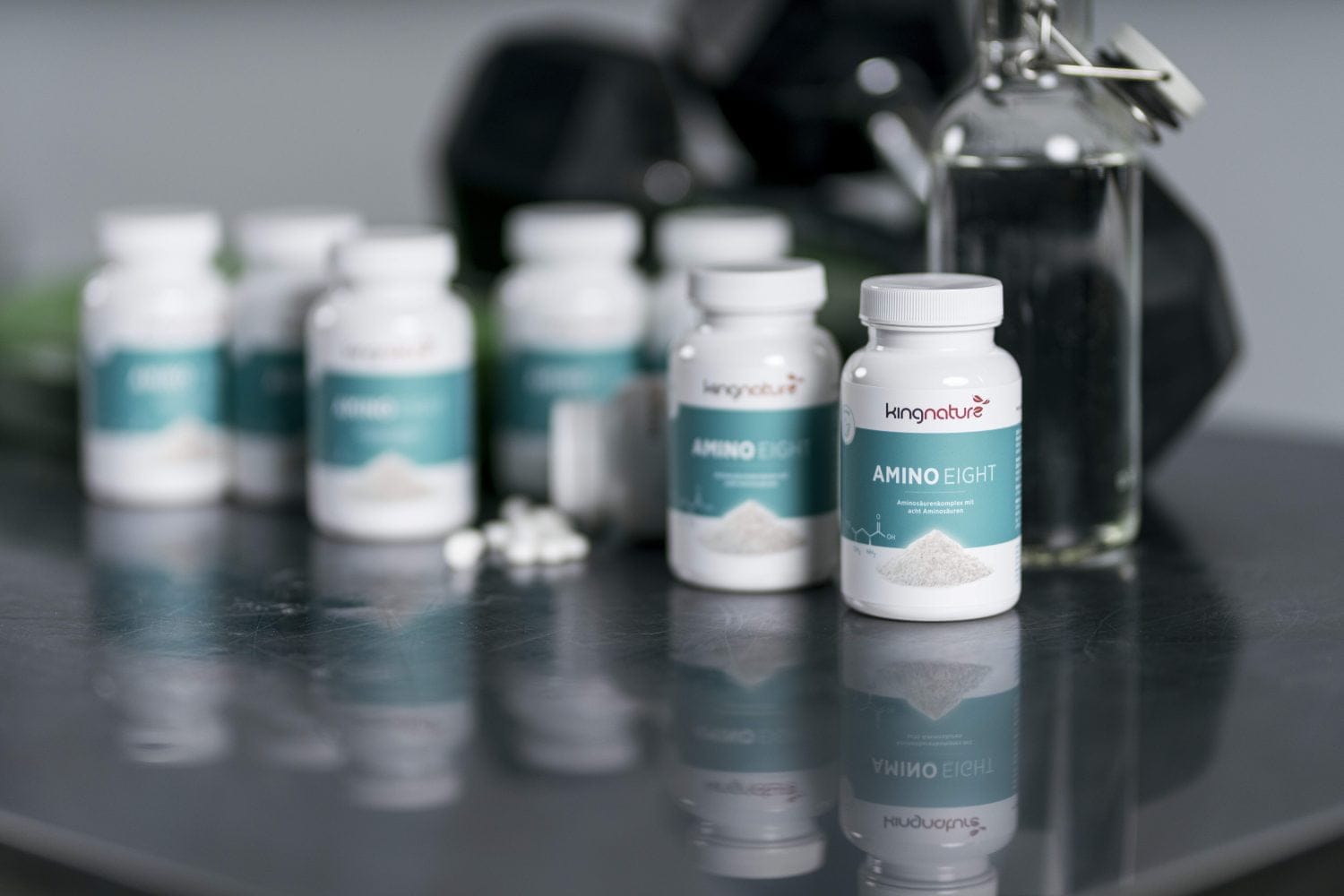 Mehrere weiße Nahrungsergänzungsmittelflaschen mit der Aufschrift „Amino Eight“ stehen auf einer reflektierenden Oberfläche, daneben einige Kapseln. Im unscharfen Hintergrund sind weitere Flaschen und eine Glasflasche mit Metallverschluss zu sehen, die Wellness und Aminosäurenunterstützung betonen.