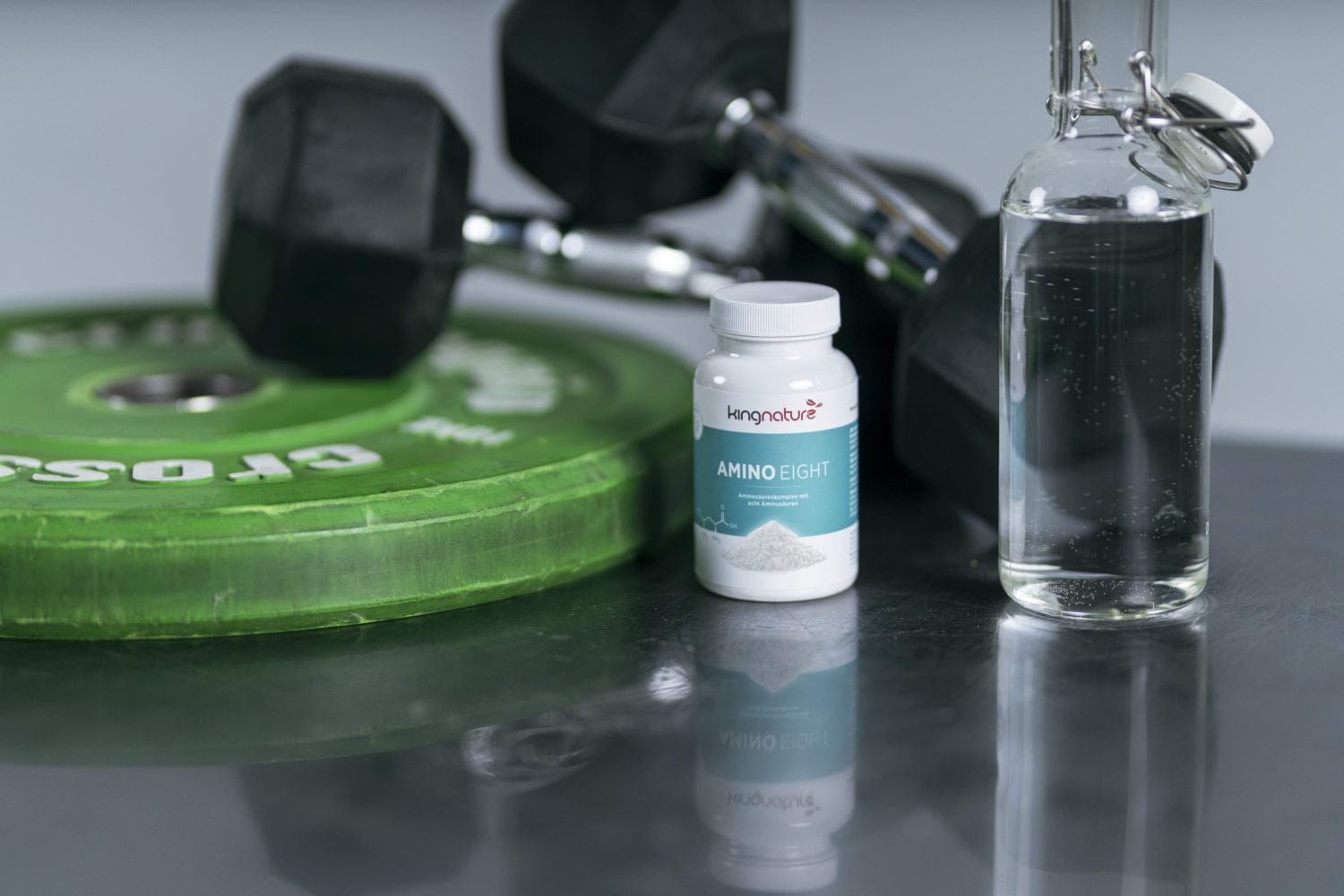 Eine Flasche mit der Aufschrift „Amino Eight“ steht auf dem Boden eines Fitnessstudios neben einer Glasflasche Wasser, einer grünen CrossFit-Hantelscheibe und zwei schwarzen Hanteln – ideal für alle, die Aminosäuren für ihr Fitnessprogramm kaufen möchten.