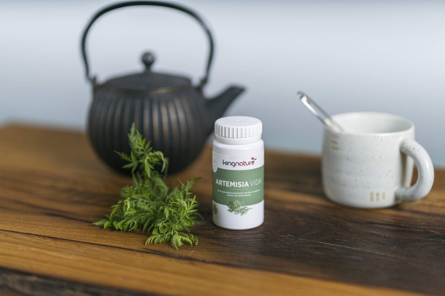 Eine weiße Flasche mit der Aufschrift „Artemisia Vida“ steht auf einem Holztisch mit grünen Blättern, neben einer schwarzen Teekanne und einer weißen Tasse mit einem Löffel – perfekt für alle, die Artemisia-Kapseln kaufen möchten.