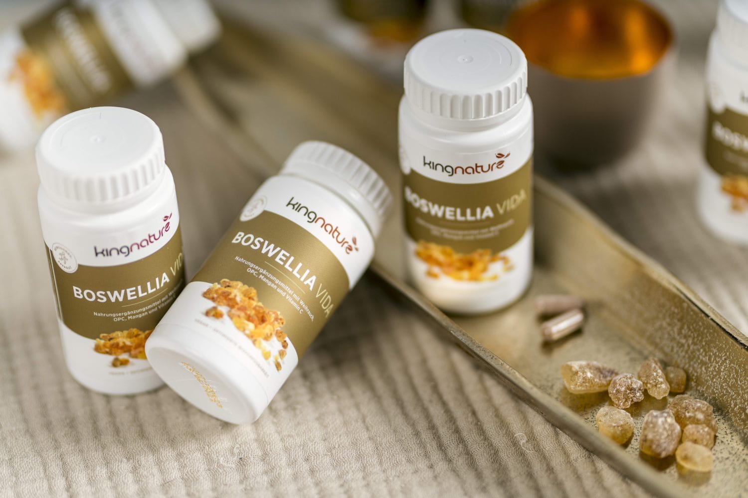 Drei Flaschen des Nahrungsergänzungsmittels Boswellia Vida von Kingnature werden auf einem Tablett neben mehreren Kapseln und Stücken aus natürlichem Boswellia-Harz präsentiert, vor einem weichen, neutral getönten Hintergrund.