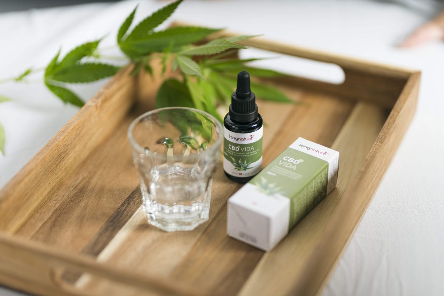 Auf einem Holztablett befinden sich eine CBD-Ölflasche mit Pipette, ein Glas Wasser, eine Produktschachtel mit der Aufschrift „CBD VIDA“ und ein Cannabisblatt, wodurch die natürlichen Elemente von CBD hervorgehoben werden.