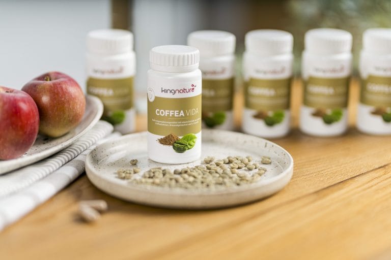 Eine Flasche Kingnature Coffea Vida-Nahrungsergänzungsmittel steht auf einem Teller mit grünen Kaffeebohnen, begleitet von roten Äpfeln, während im Hintergrund weitere Flaschen verschwommen erscheinen.