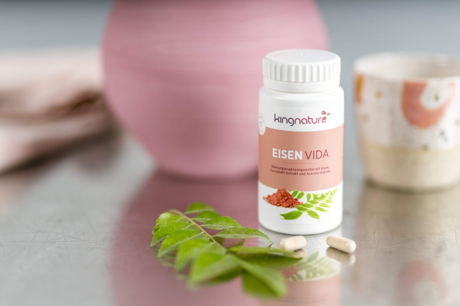 Eine Flasche mit der Aufschrift „kingnature EISEN VIDA“ steht zusammen mit zwei Kapseln, einem grünen Blatt, einer rosa Vase und einer Keramiktasse in einer sanften, natürlichen Umgebung auf einem Tisch – ideal für alle, die Eisen Vida kaufen möchten.