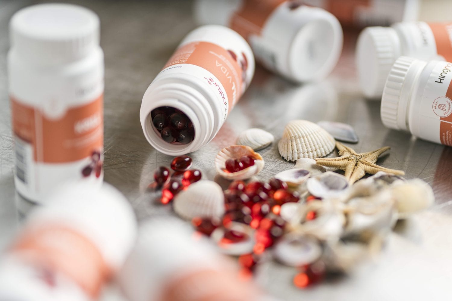 Aus mehreren weißen Flaschen mit orangefarbenen Etiketten, einige davon offen und umgekippt, ergießen sich rote Kapseln auf eine Oberfläche, auf der Muscheln und ein kleiner Seestern verstreut sind.