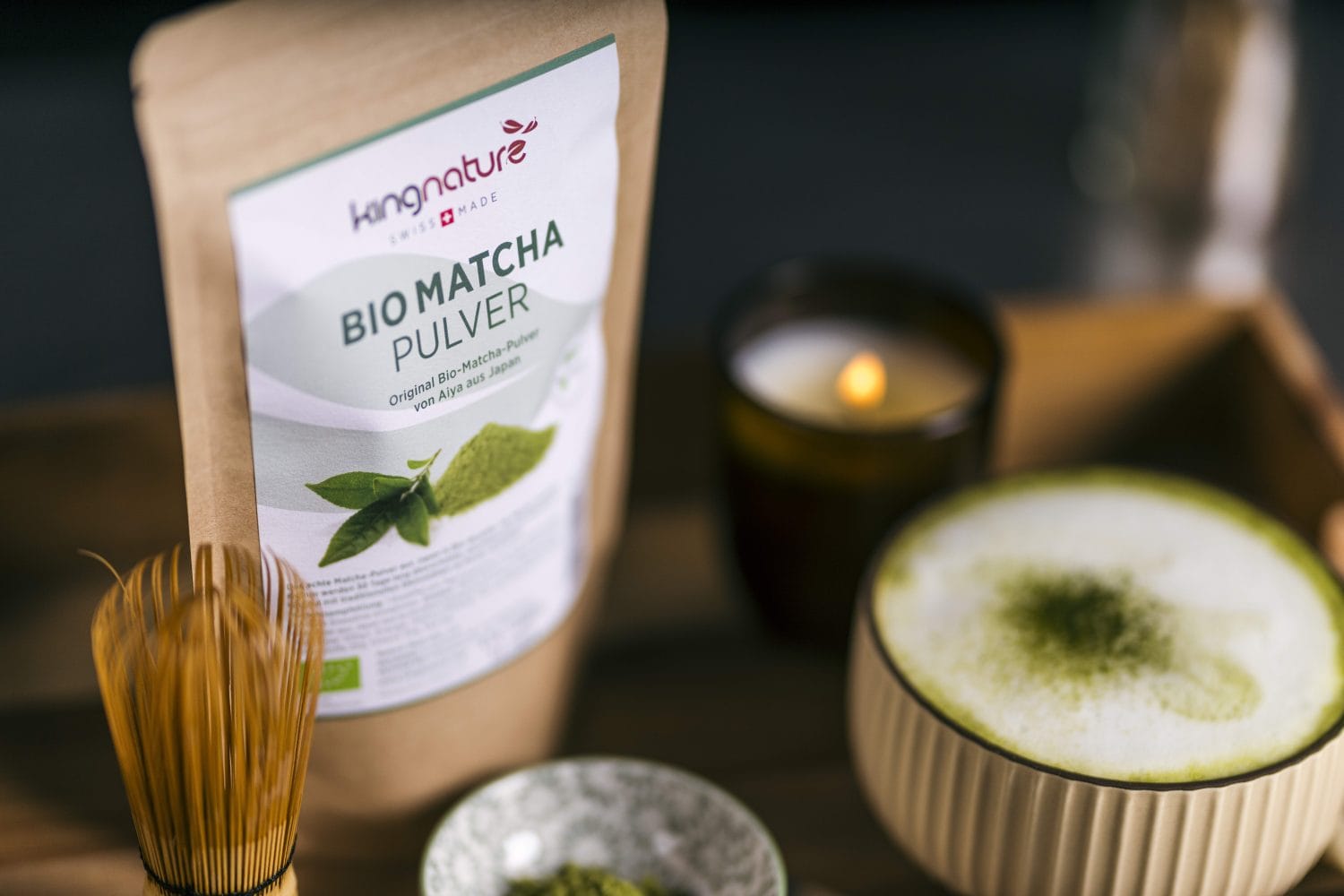 Eine Packung Bio Matcha Pulver steht auf einem Tablett neben einer Schale Matcha-Getränk, einem kleinen Schälchen Pulver, einem Bambusbesen und einer brennenden Kerze. Die gemütliche Szene lädt dazu ein, Matcha Pulver zu kaufen und die warme Atmosphäre zu genießen.