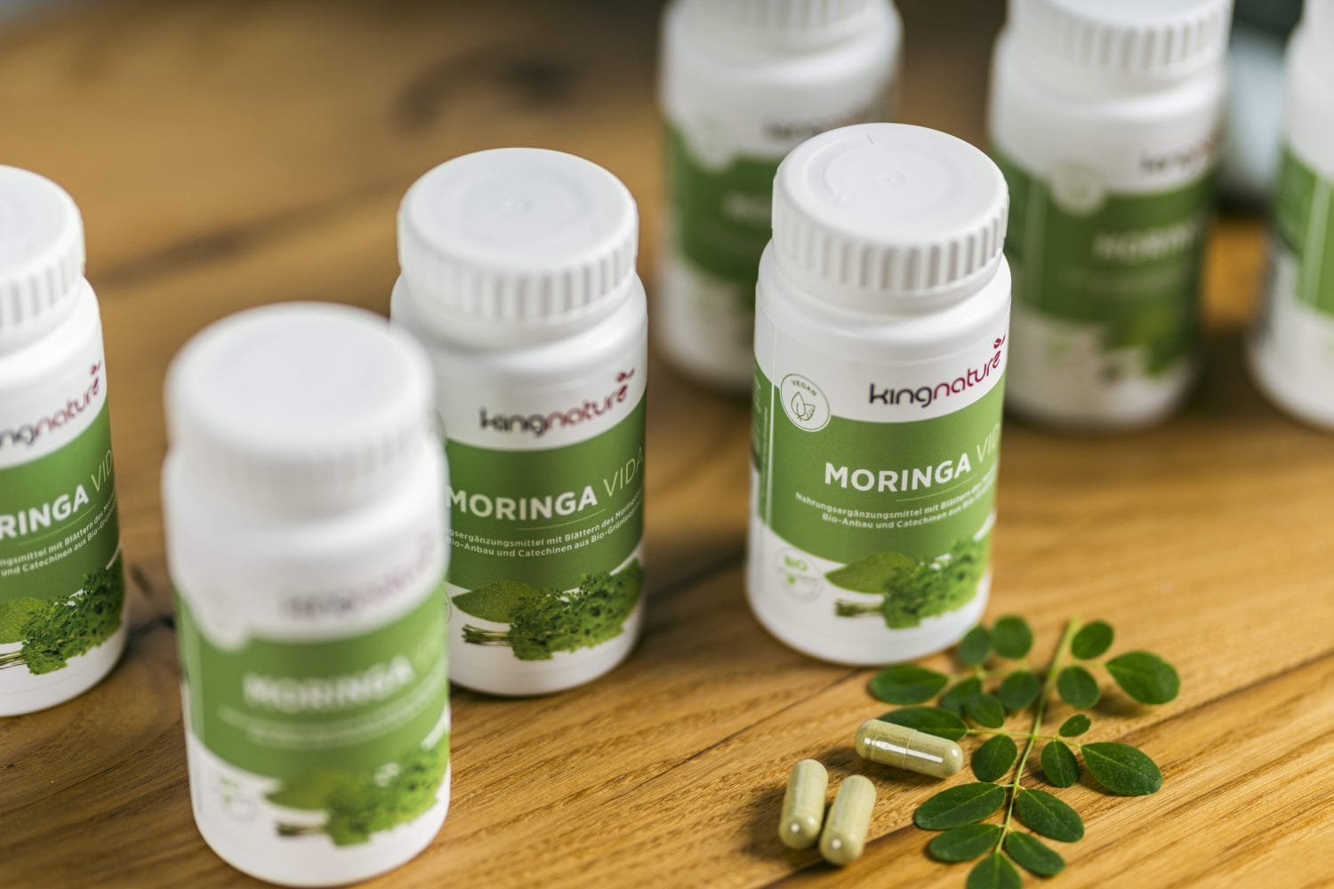 Mehrere weiße Flaschen mit der Aufschrift „Moringa Vida“ von kingnature sind auf einer Holzoberfläche angeordnet. Neben den Flaschen heben zwei grüne Kapseln und frische Moringablätter die natürlichen Vorteile von Moringa hervor.