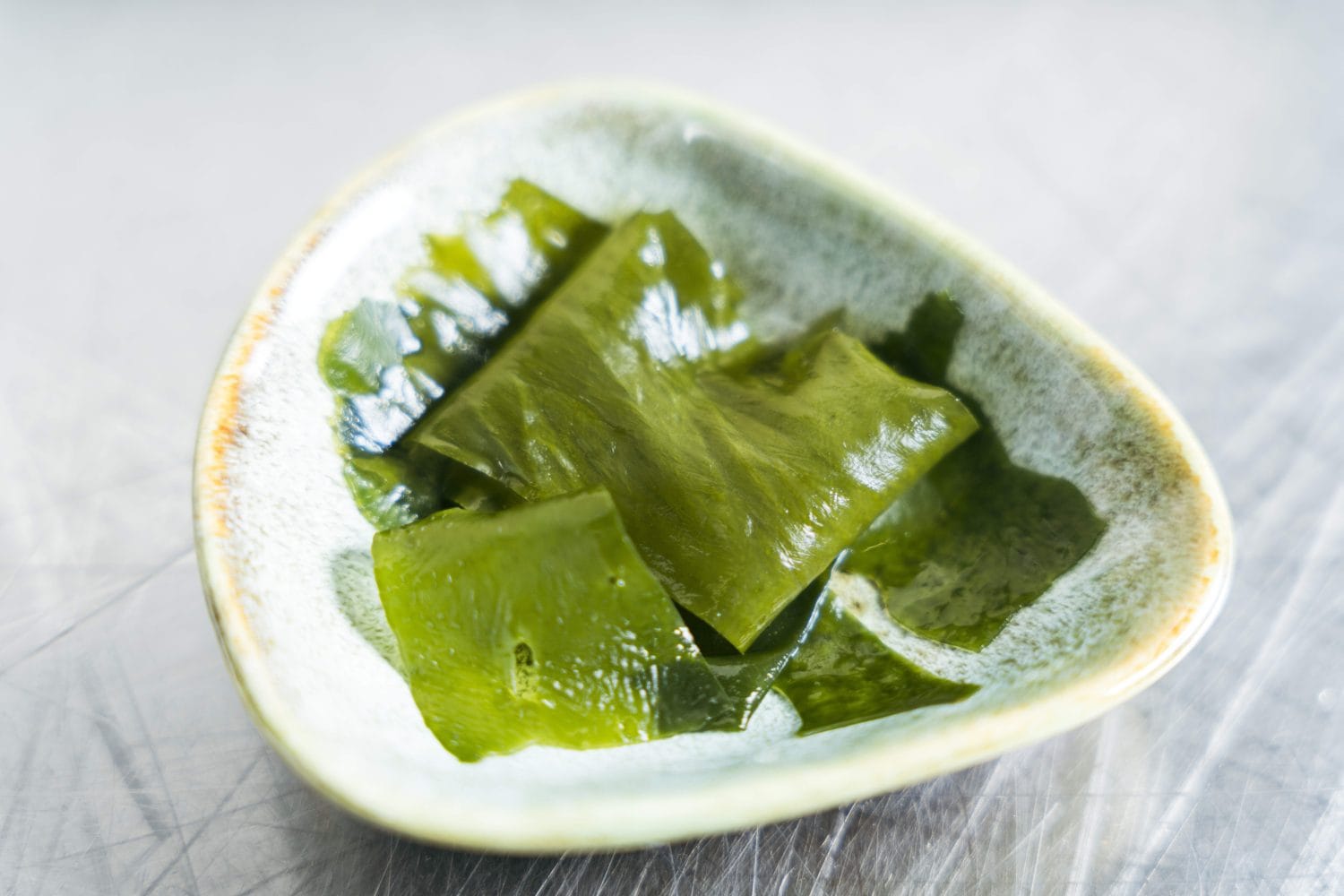 Auf einer hellgrauen Oberfläche liegt eine kleine Keramikschale mit mehreren glänzenden, grünen Algenscheiben – natürlich reich an veganem Omega-3-Öl.