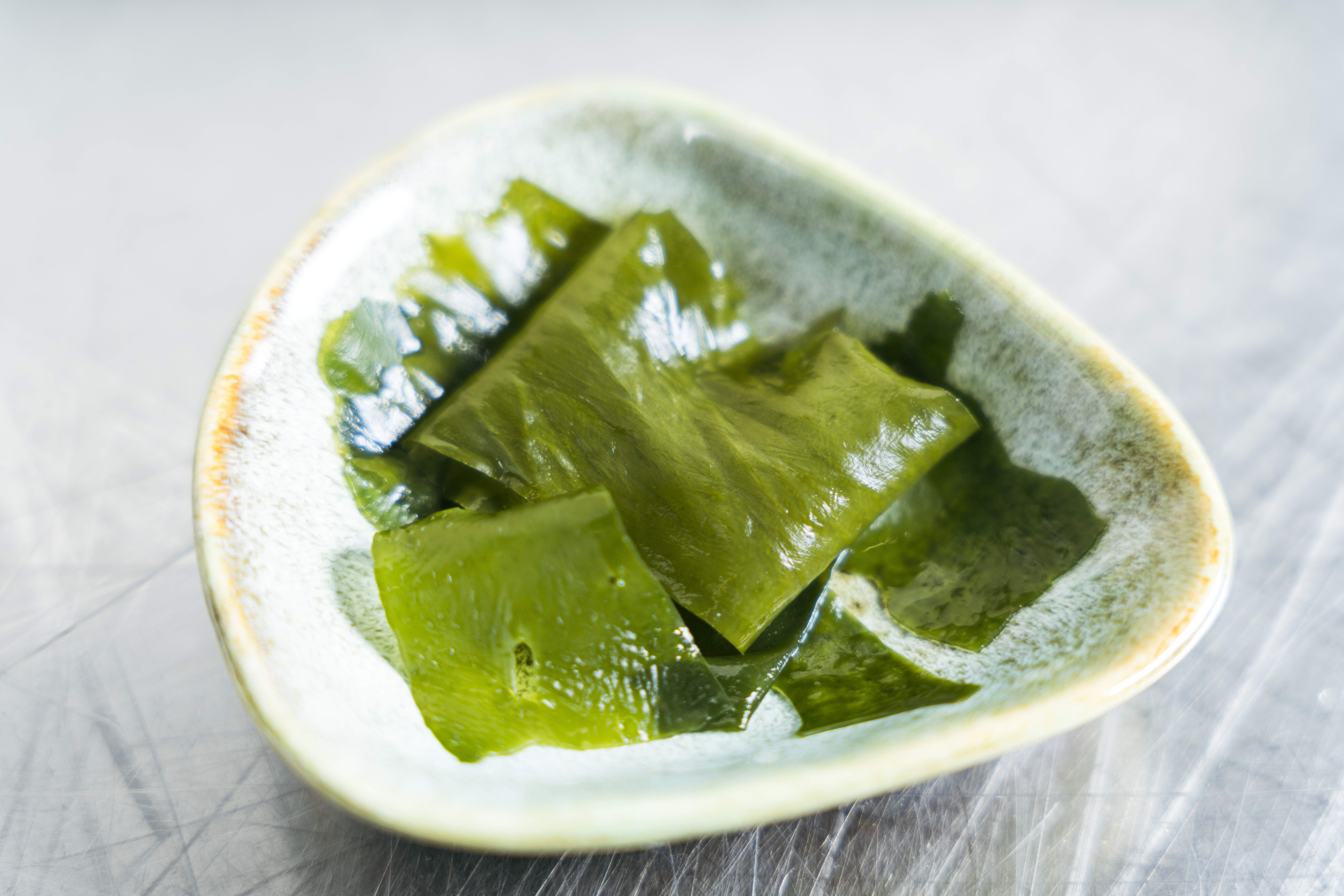 Auf einer hellgrauen Oberfläche liegt eine kleine Keramikschale mit mehreren glänzenden, grünen Algenscheiben – natürlich reich an veganem Omega-3-Öl.