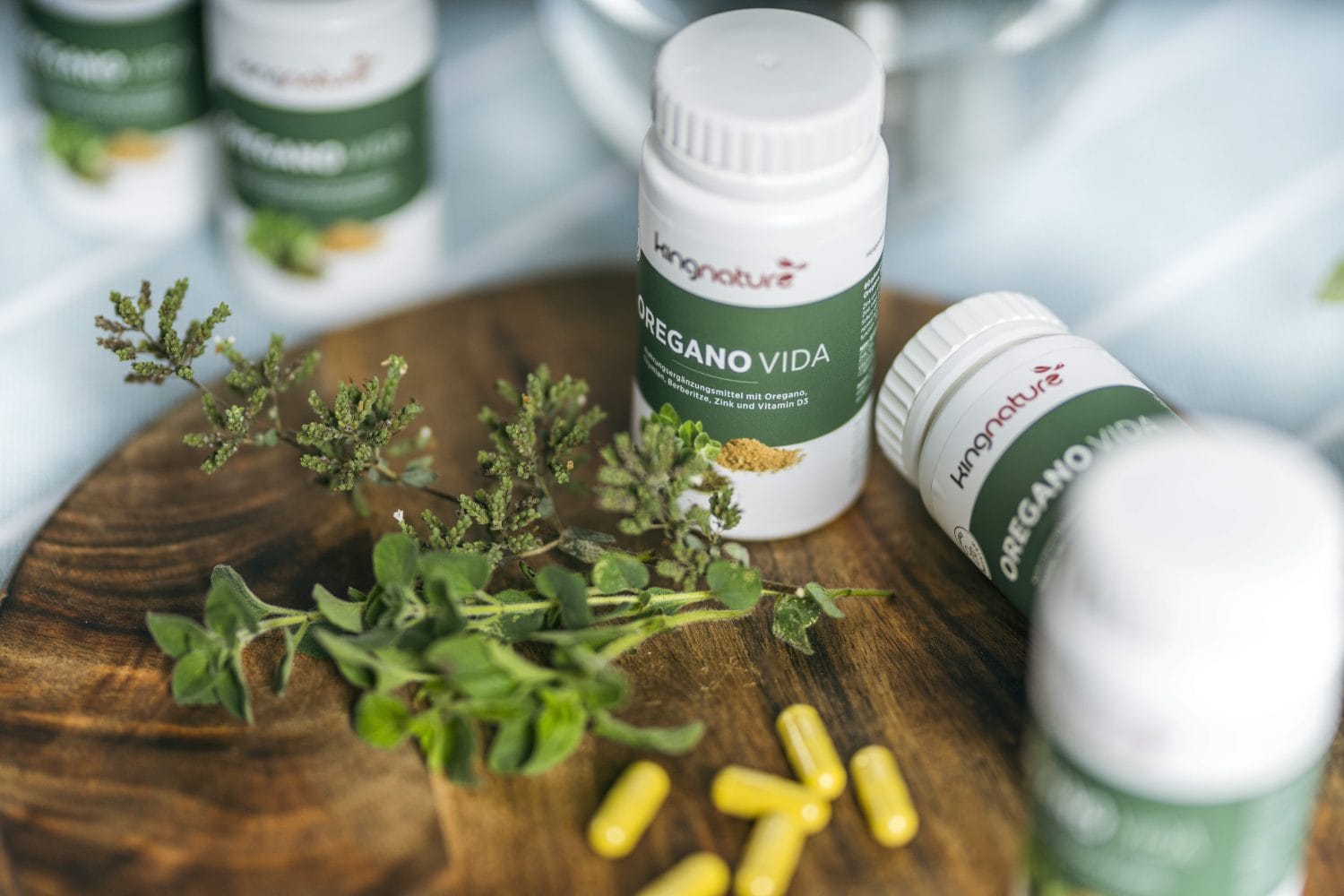 Flaschen mit der Aufschrift „Oregano Vida“ stehen neben Oreganozweigen und gelben Kapseln auf einer Holzfläche und machen auf das natürliche Nahrungsergänzungsmittel aufmerksam. Einige Flaschen stehen aufrecht, eine liegt auf der Seite.
