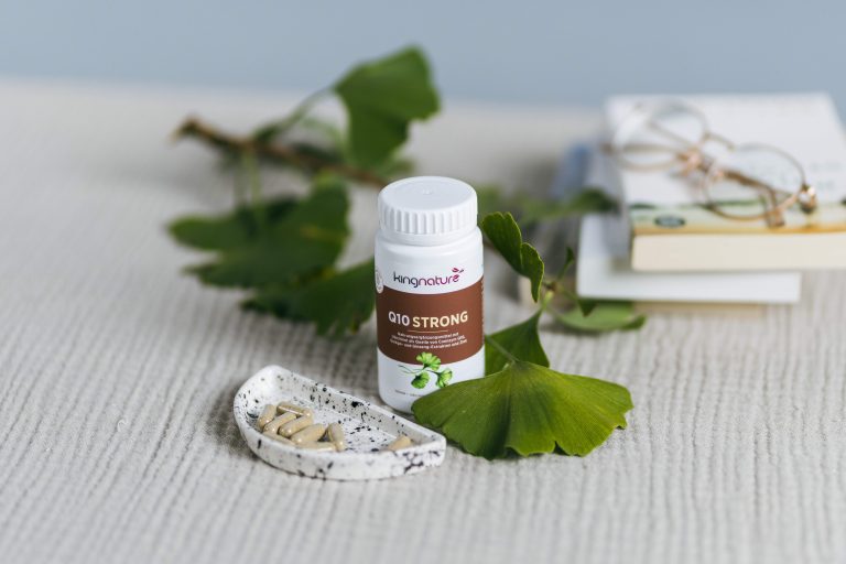 Eine Flasche Q10 Strong-Nahrungsergänzungsmittel, ein Energiepaket Medium, steht auf einer strukturierten Oberfläche neben einer Keramikschale mit Kapseln, grünen Ginkgoblättern, einer Brille und einem Stapel Bücher im Hintergrund.