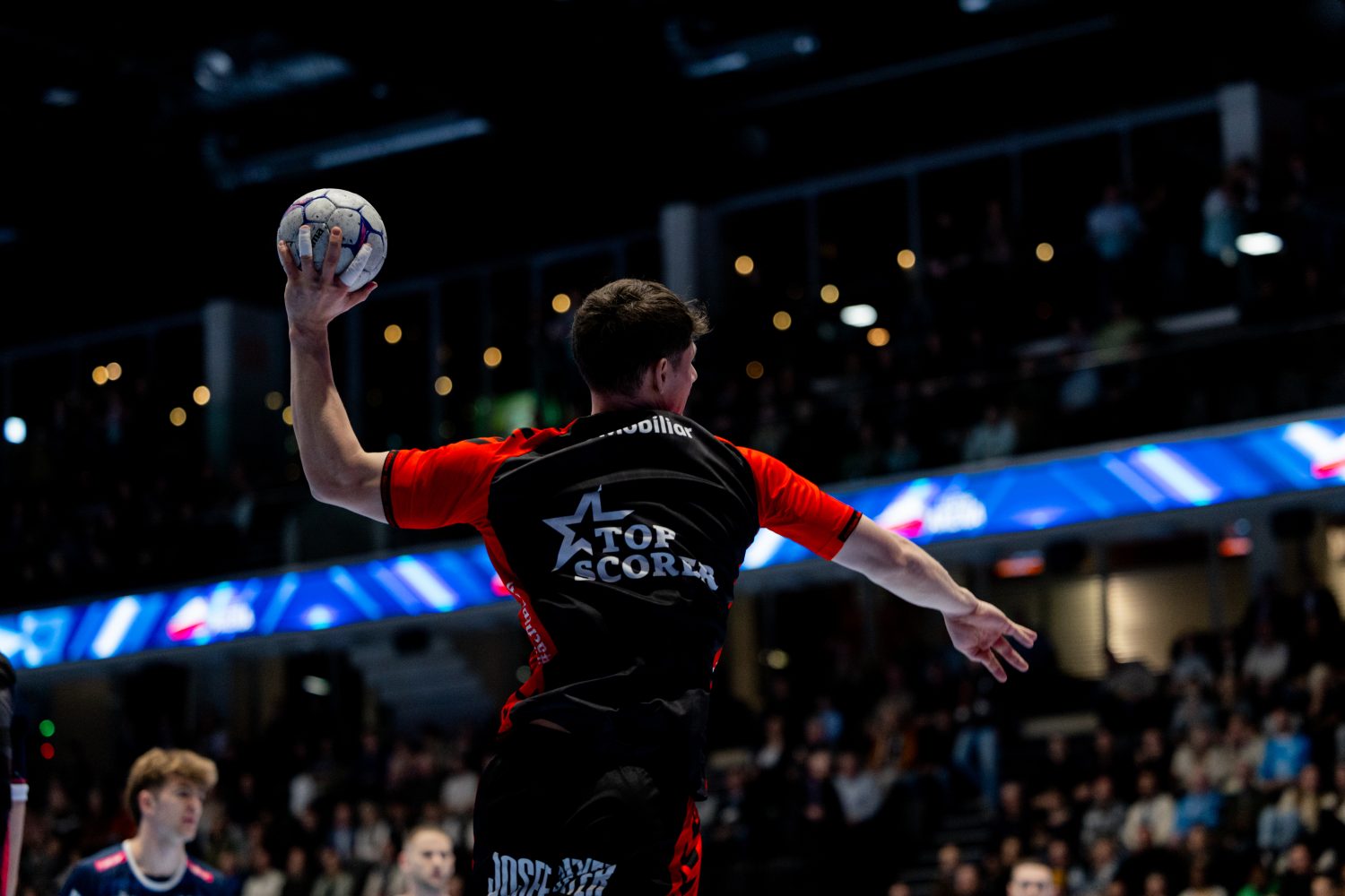 Levin Wanner, in einem schwarz-roten Trikot mit der Aufschrift "Top Scorer" (Torschützenkönig), bereitet sich während eines Hallenhandballspiels auf den Wurf vor, während das Publikum im Hintergrund unscharf zusieht.