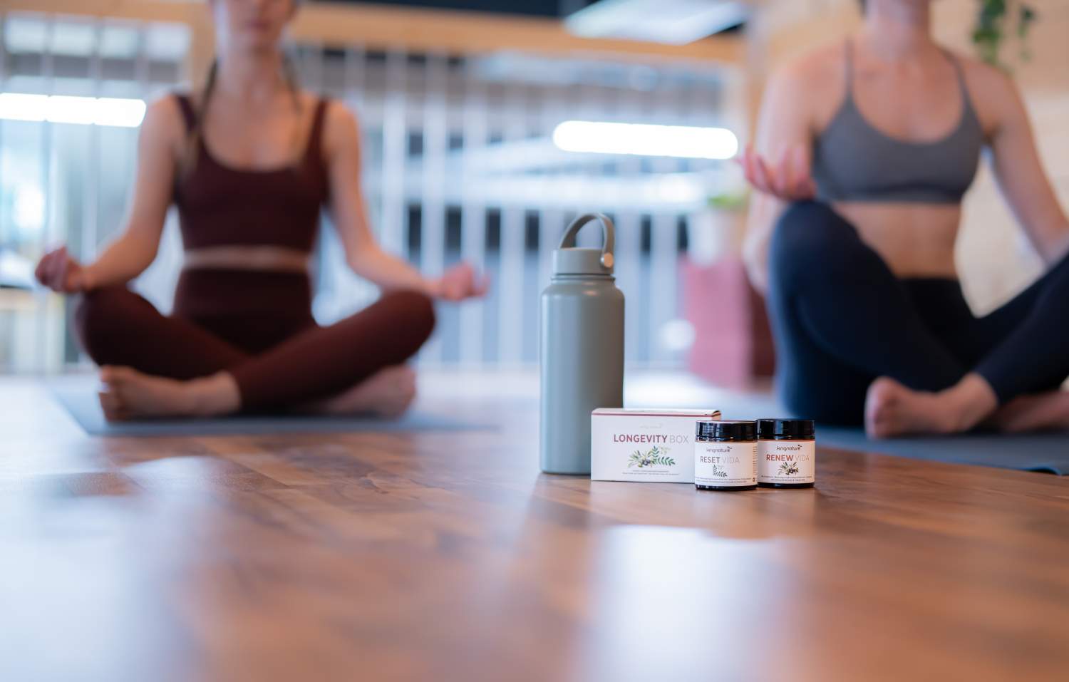 Zwei Personen in Sportkleidung sitzen im Schneidersitz auf Yogamatten und meditieren im Innenbereich. Im Vordergrund sind eine Wasserflasche und zwei Wellness-Nahrungsergänzungsmittel auf einem Holzboden zu sehen.