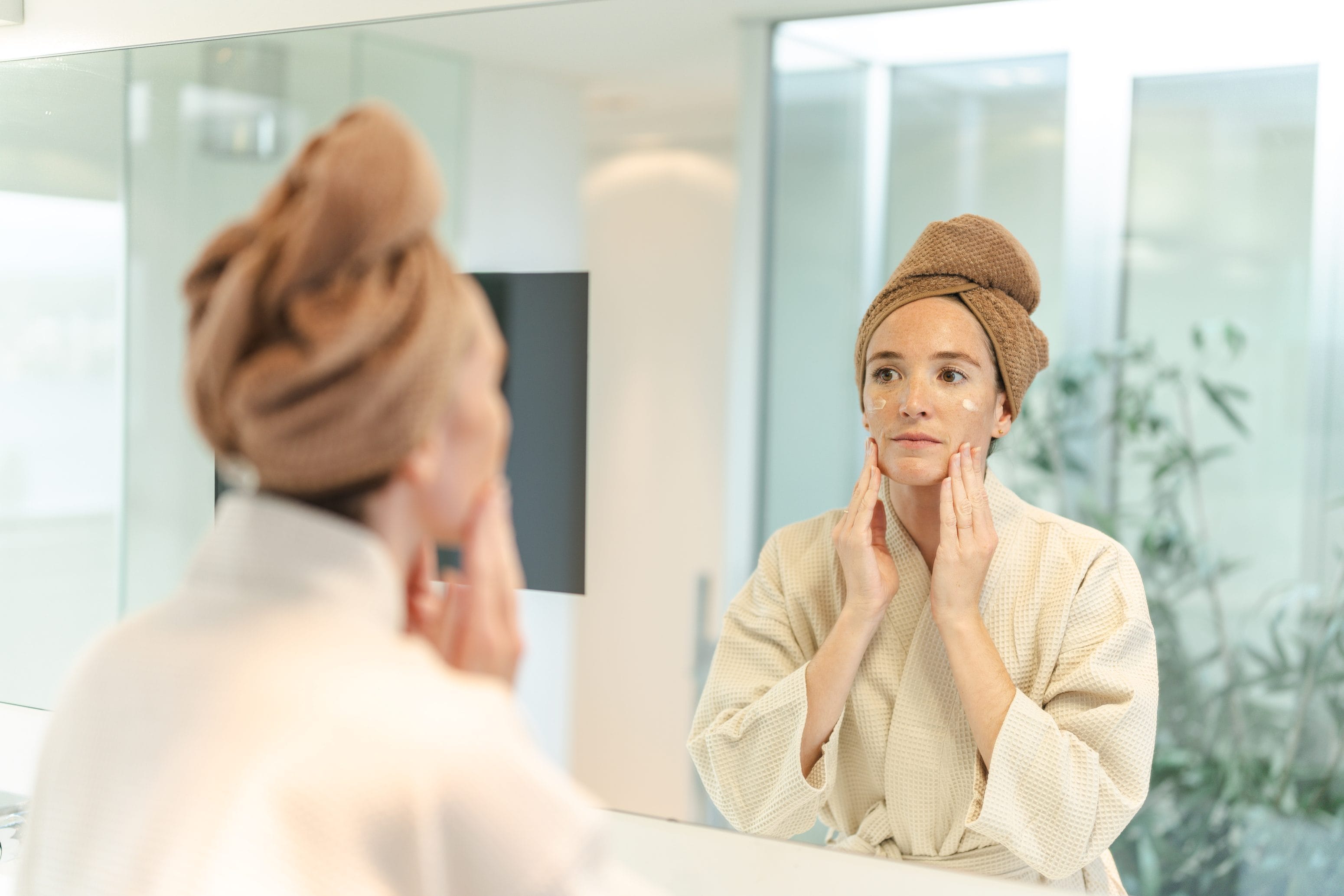 Eine Frau im Bademantel mit einem Handtuch um den Kopf trägt Feuchtigkeitscreme auf ihr Gesicht auf, während sie sich im Badezimmerspiegel betrachtet.