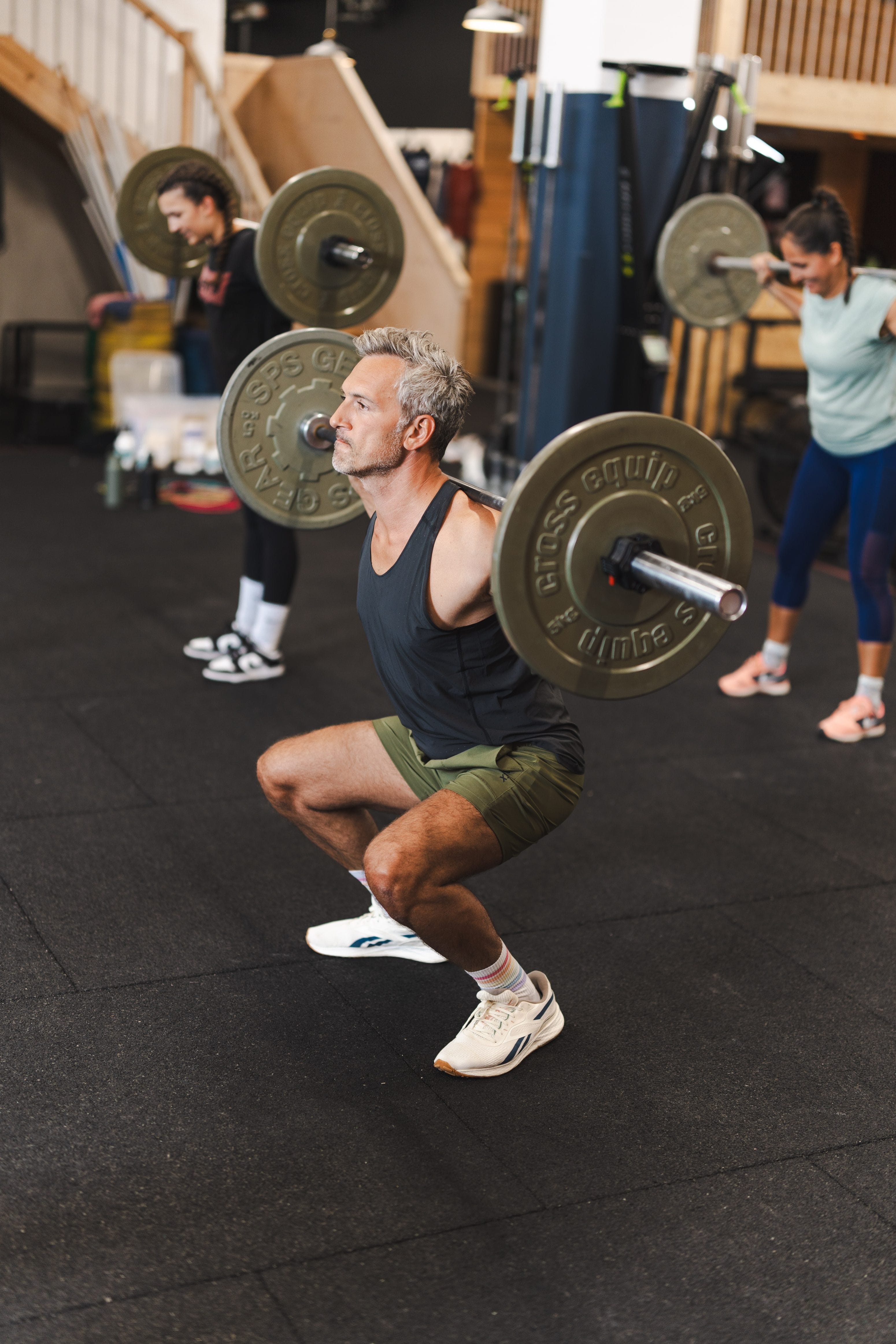 Ein Mann in Sportkleidung macht in einem Fitnessstudio Kniebeugen mit der Langhantel, während im Hintergrund zwei Personen Gewichte stemmen. Alle konzentrieren sich auf ihr Krafttraining in einem gut ausgestatteten Indoor-Trainingsbereich.