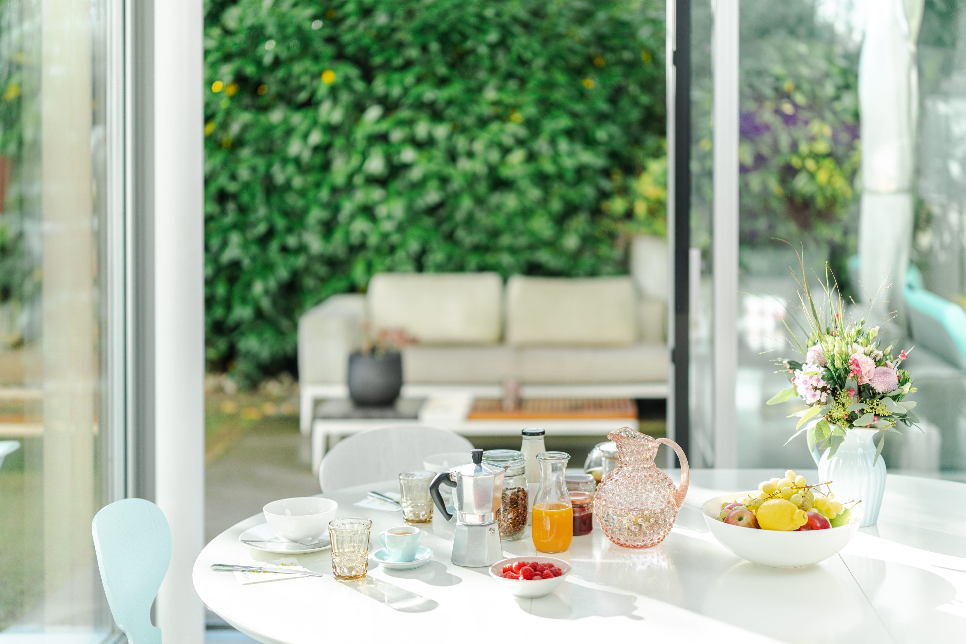 Ein heller, sonnendurchfluteter Esstisch, gedeckt zum Frühstück mit Saft, Obst, Kaffee und Gebäck. Eine Glastür öffnet sich zu einem Garten mit einem gemütlichen Sofa und üppigem Grün im Hintergrund. Ein Strauß frischer Blumen verleiht der Szenerie Charme.