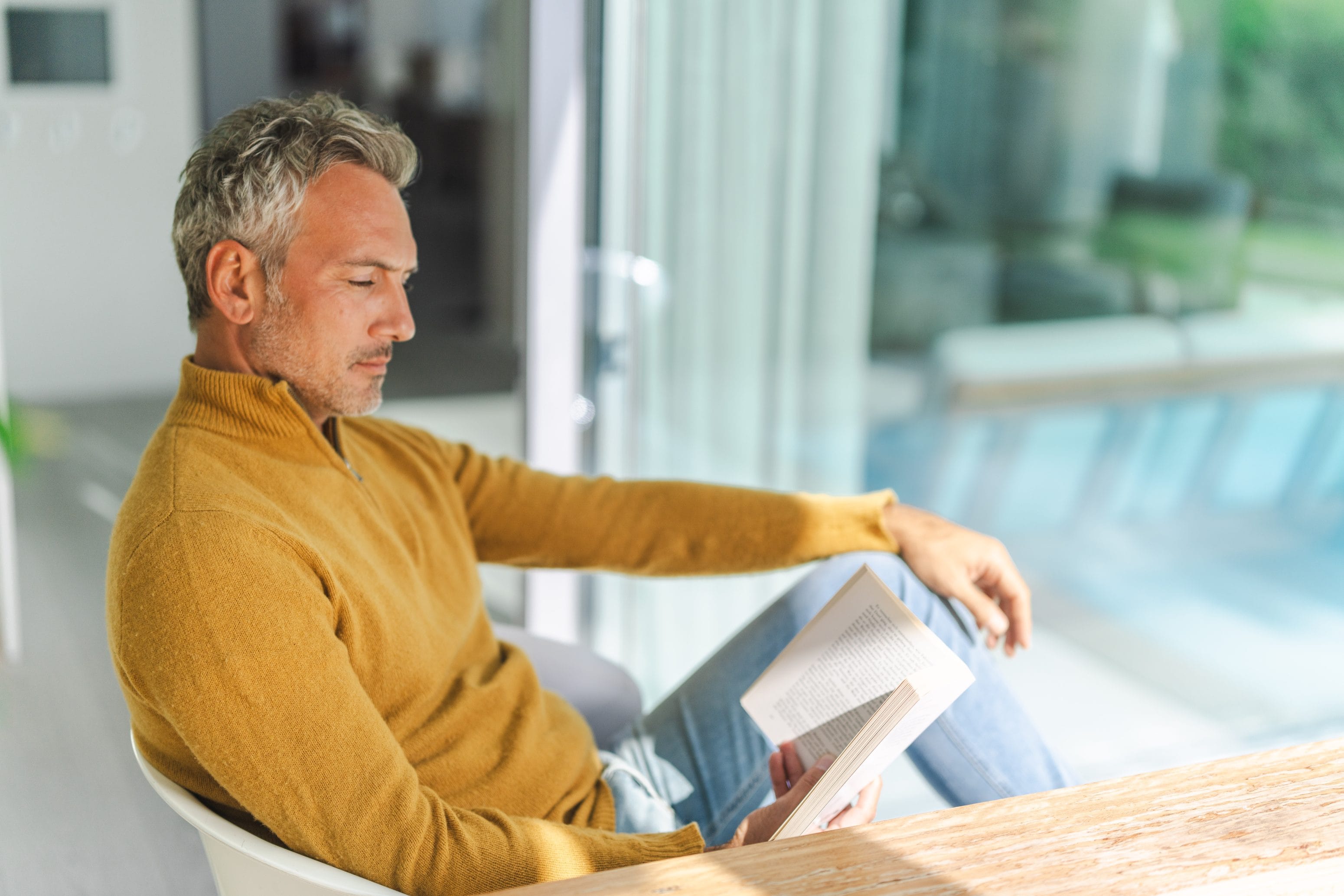 Ein Mann mit grauem Haar und einem gelben Pullover sitzt am Fenster und liest ein Buch. Sonnenlicht fällt herein und sorgt für eine friedliche und entspannte Stimmung.