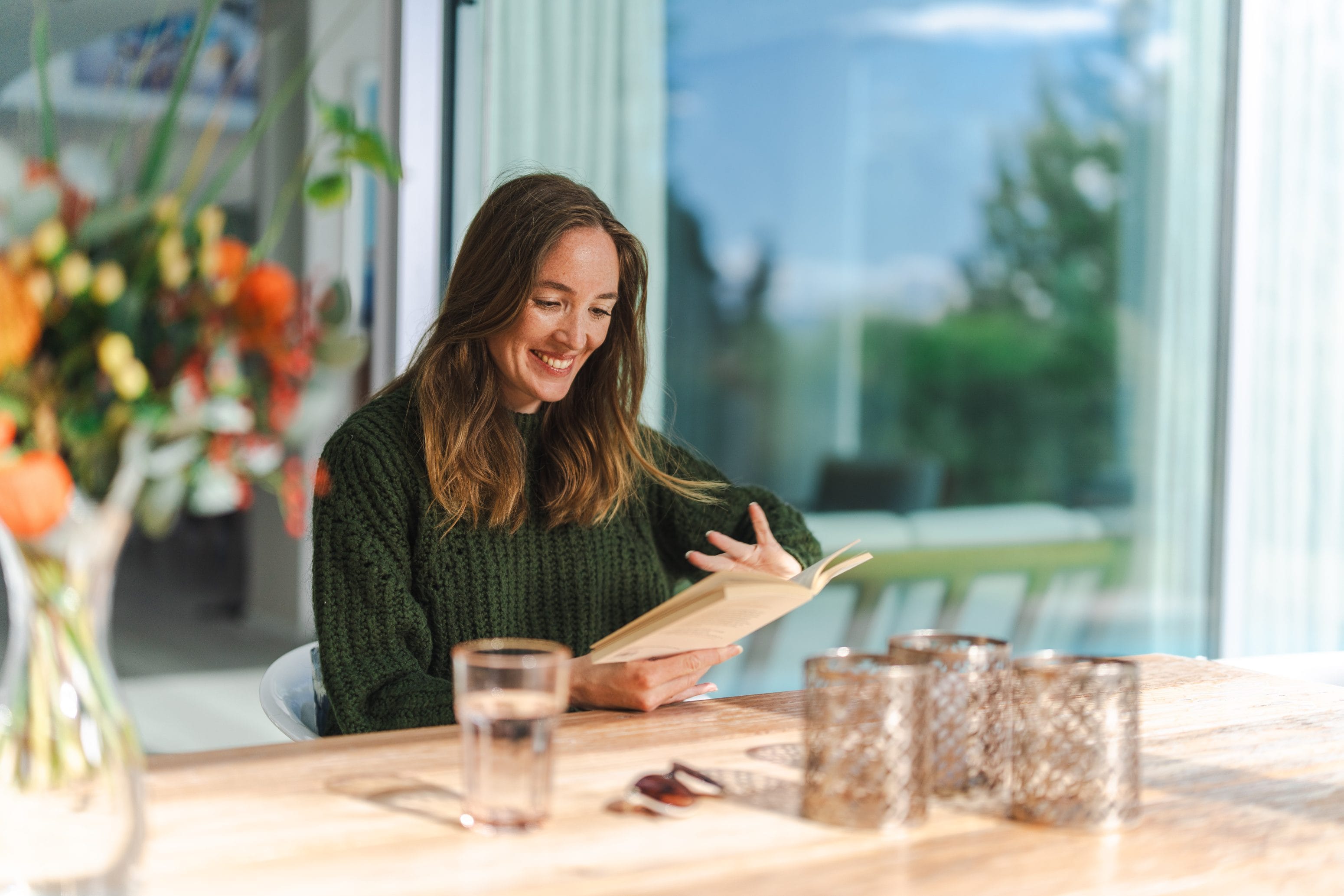 Eine Frau mit langen braunen Haaren und einem grünen Pullover sitzt lächelnd an einem Holztisch, liest ein Buch. Auf dem Tisch stehen ein Glas Wasser, Kerzenständer und eine Vase mit frischen Blumen. Sonnenlicht fällt durch die großen Fenster und schafft eine gemütliche Atmosphäre.
