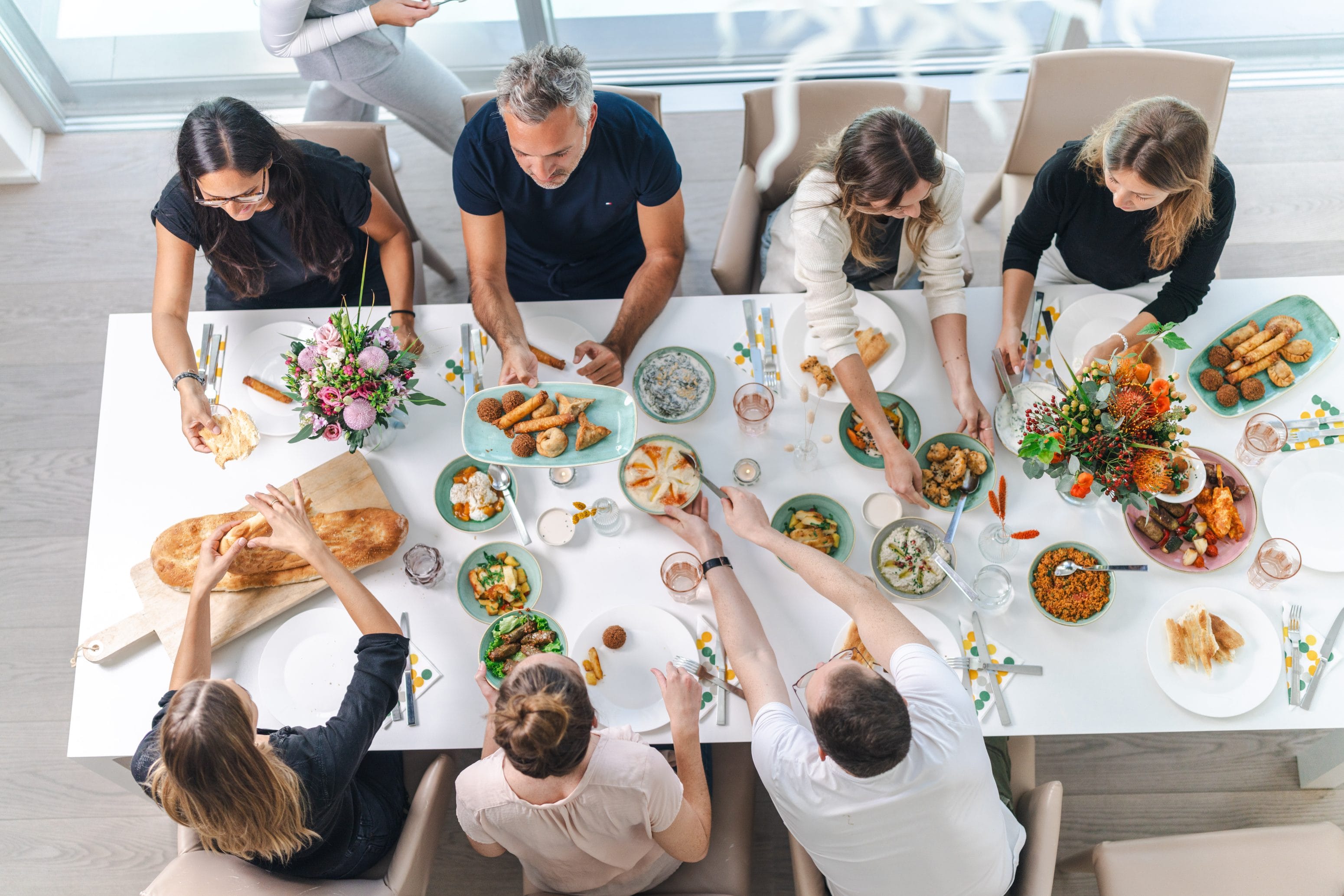 Eine Gruppe von Menschen sitzt um einen Tisch und genießt gemeinsam frische Salate und Vorspeisen. Der Tisch ist mit Blumen dekoriert und mit mediterranen Gerichten gefüllt, was eine lebendige, gemeinschaftliche Atmosphäre schafft.