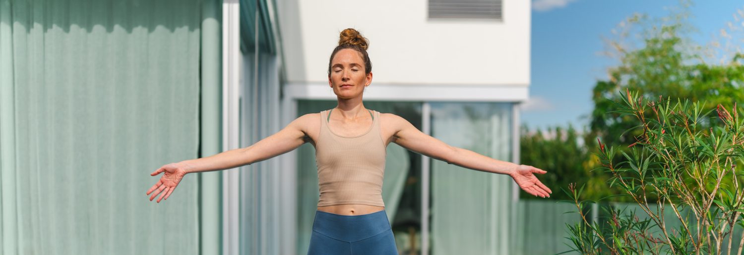 Eine Frau steht mit ausgestreckten Armen auf einer Yogamatte neben einem Pool im Freien in der Nähe eines modernen Hauses. Sie trägt ein beiges Tanktop und blaue Leggings, hat die Augen geschlossen und übt Achtsamkeit in einer friedlichen Umgebung.