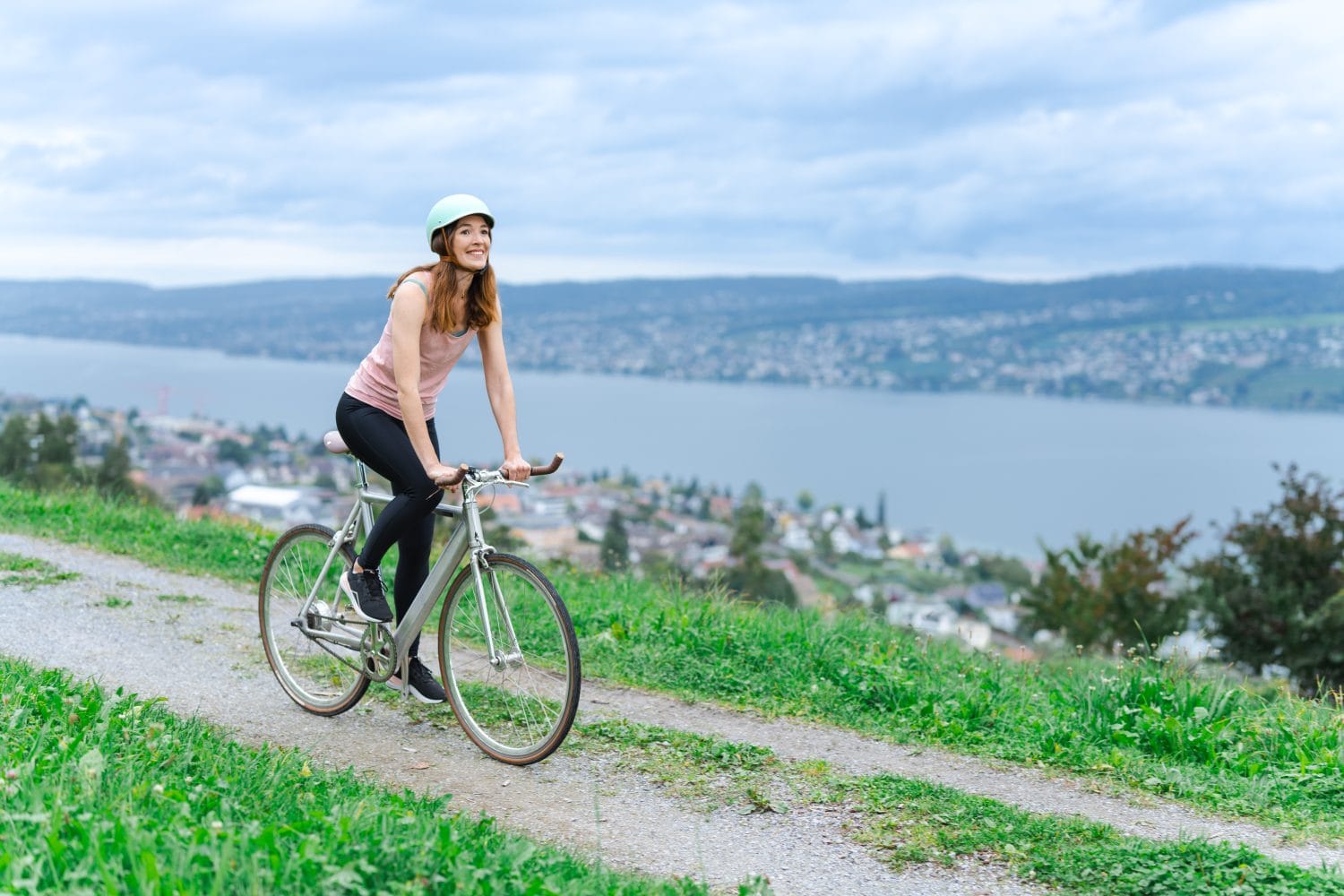 Eine Frau mit Helm fährt fröhlich mit einem E-Bike auf einem grasbewachsenen Hügelpfad. Im Hintergrund erstrecken sich unter einem bewölkten Himmel ein großer See, eine Stadt und Hügel.