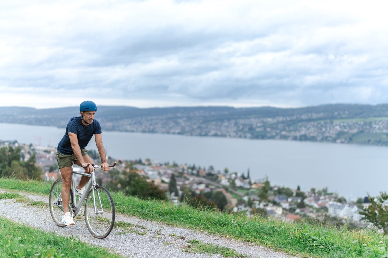 Ein Mann mit blauem Helm fährt mit dem Fahrrad auf einem Schotterweg oberhalb einer Stadt, im Hintergrund ein großer See und sanfte Hügel unter einem bewölkten Himmel, und genießt das malerische Abenteuer im Freien.