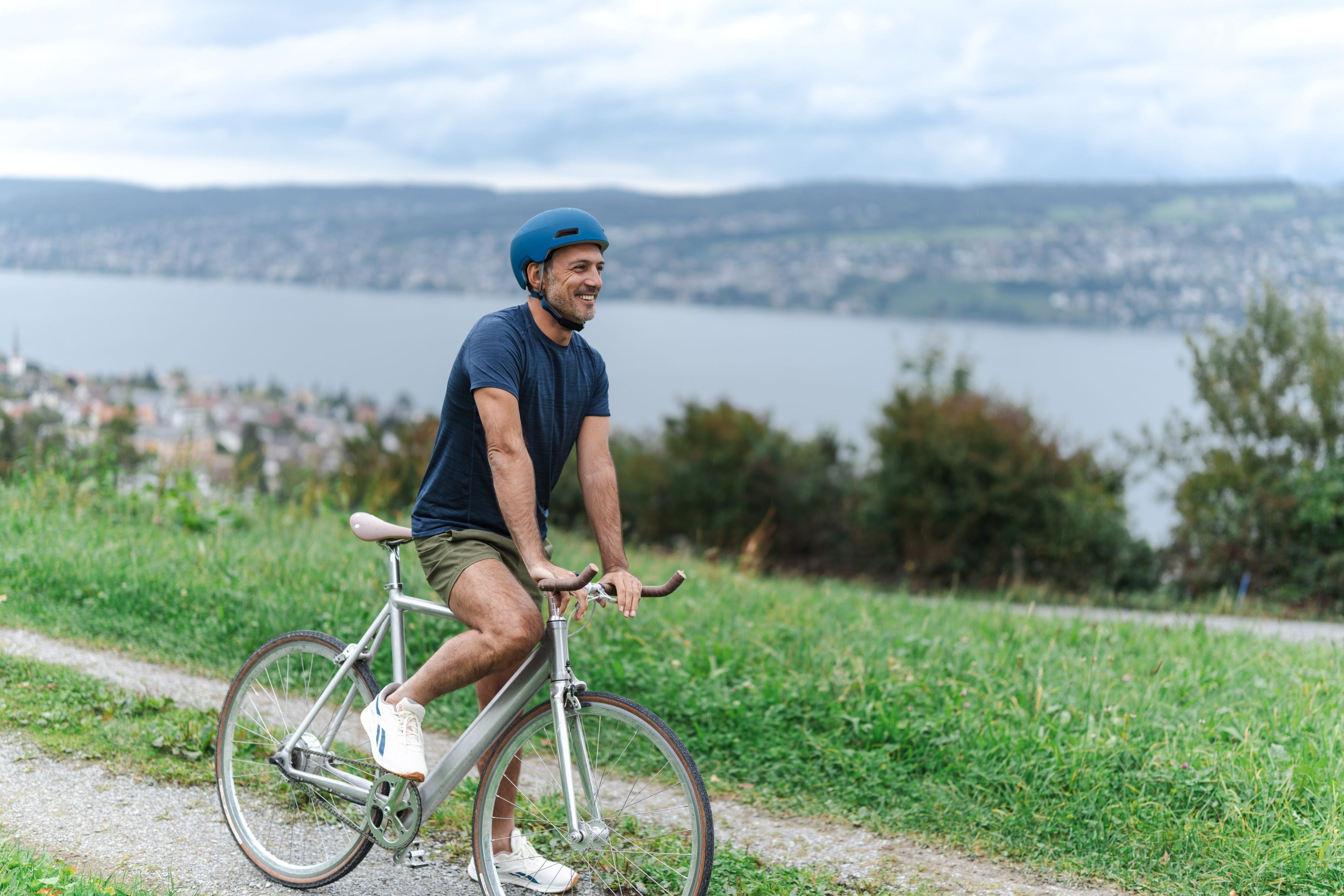 Ein Mann mit blauem Helm, marineblauem T-Shirt und Shorts fährt mit dem Fahrrad auf einem Weg mit Blick auf einen See und eine Stadt, im Hintergrund üppig grüne Hügel und einen bewölkten Himmel.
