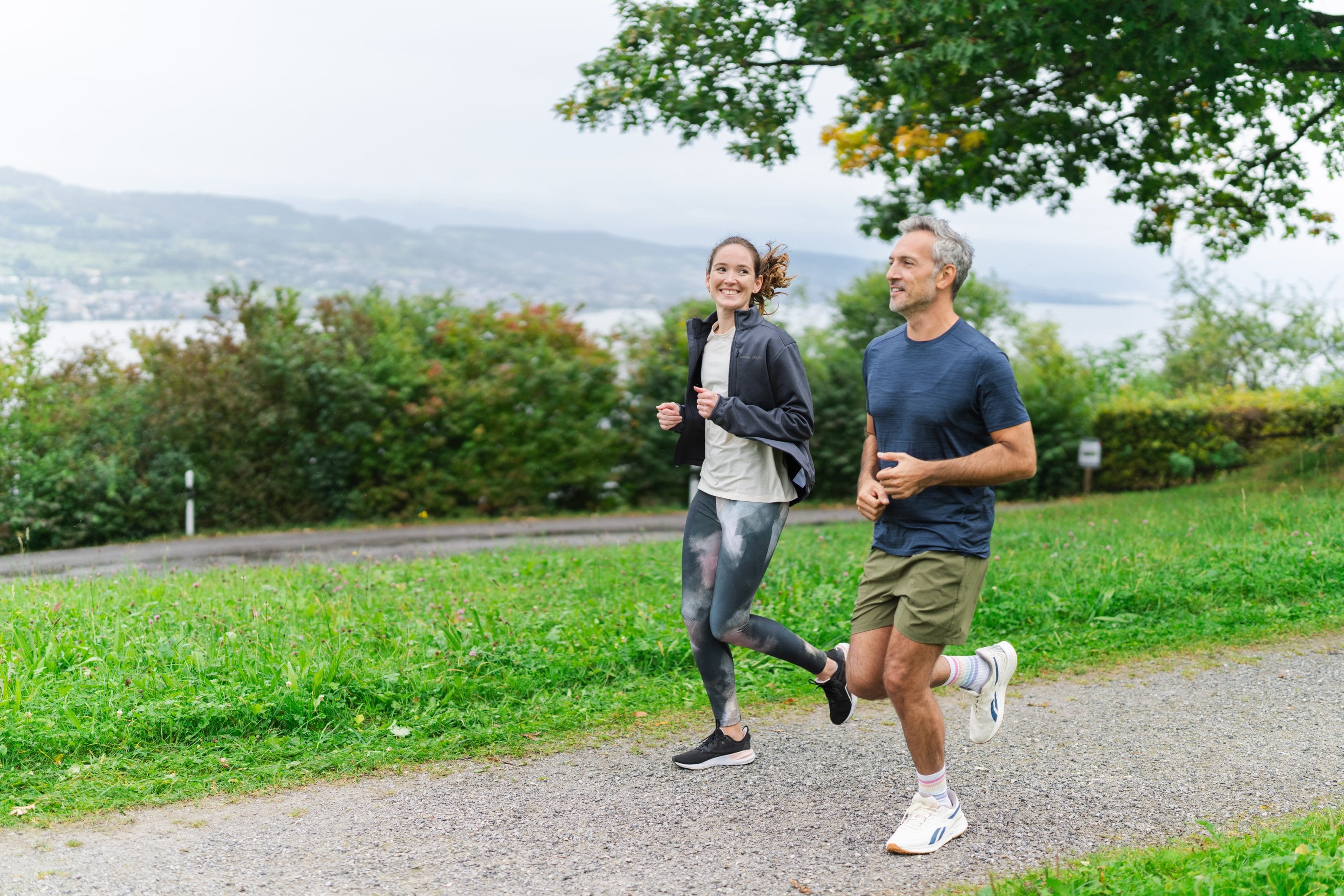 Une femme et un homme font leur jogging ensemble sur un chemin de gravier dans un parc verdoyant, tout en souriant. Entourés d'arbres et de buissons, ils profitent du calme de la nature avec des vues pittoresques de collines et d'eau scintillante en arrière-plan.