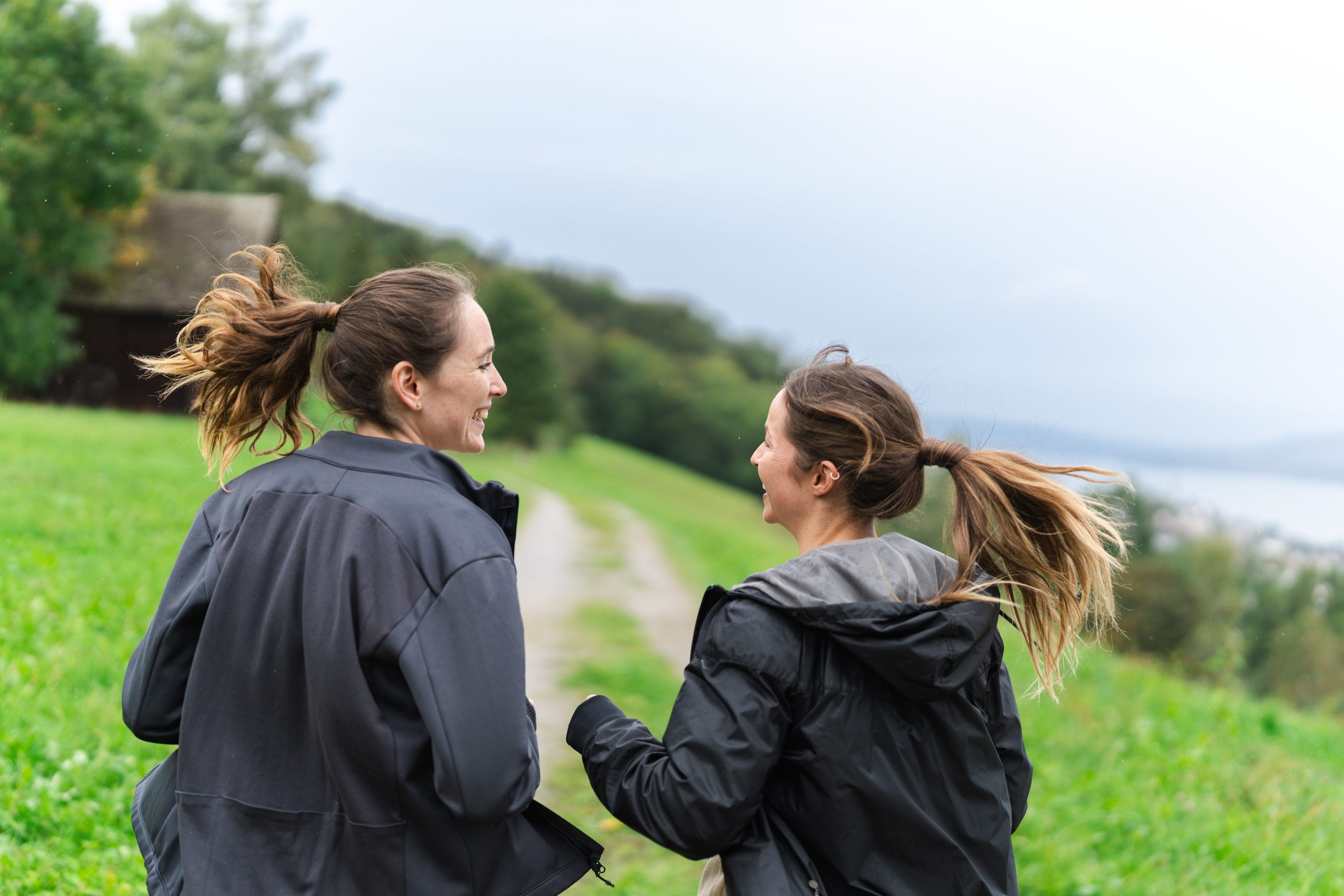 Zwei Frauen mit Pferdeschwänzen und dunklen Jacken lächeln sich an, während sie unter einem bewölkten Himmel auf einem von Bäumen gesäumten Graspfad spazieren und die frische Luft genießen.