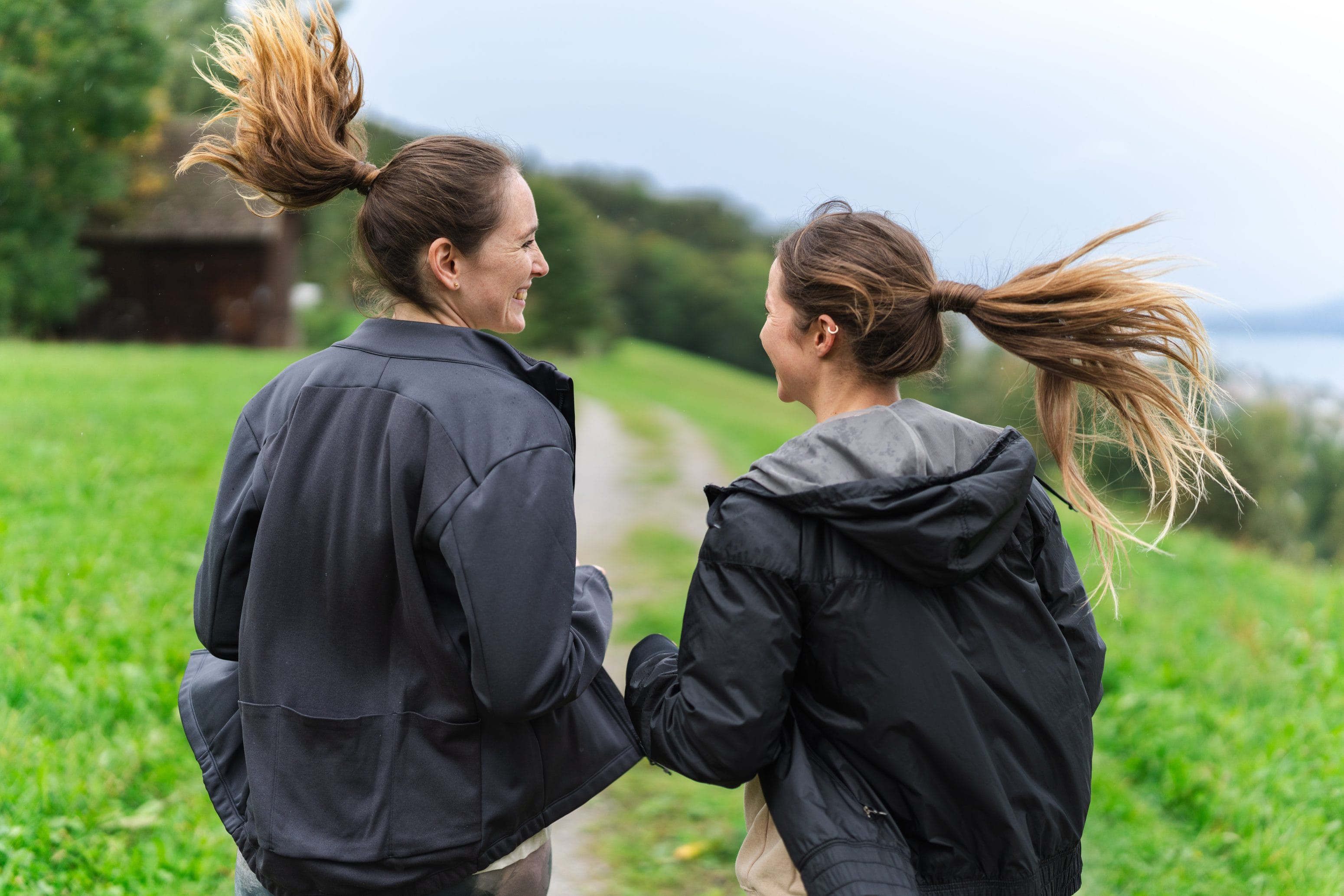 Two women with ponytails are jogging side by side on a grassy path and smiling at each other. They’re wearing jackets and are out enjoying the fresh air and the greenery—perfect for an energetic start to the day, rich in vitamin C.