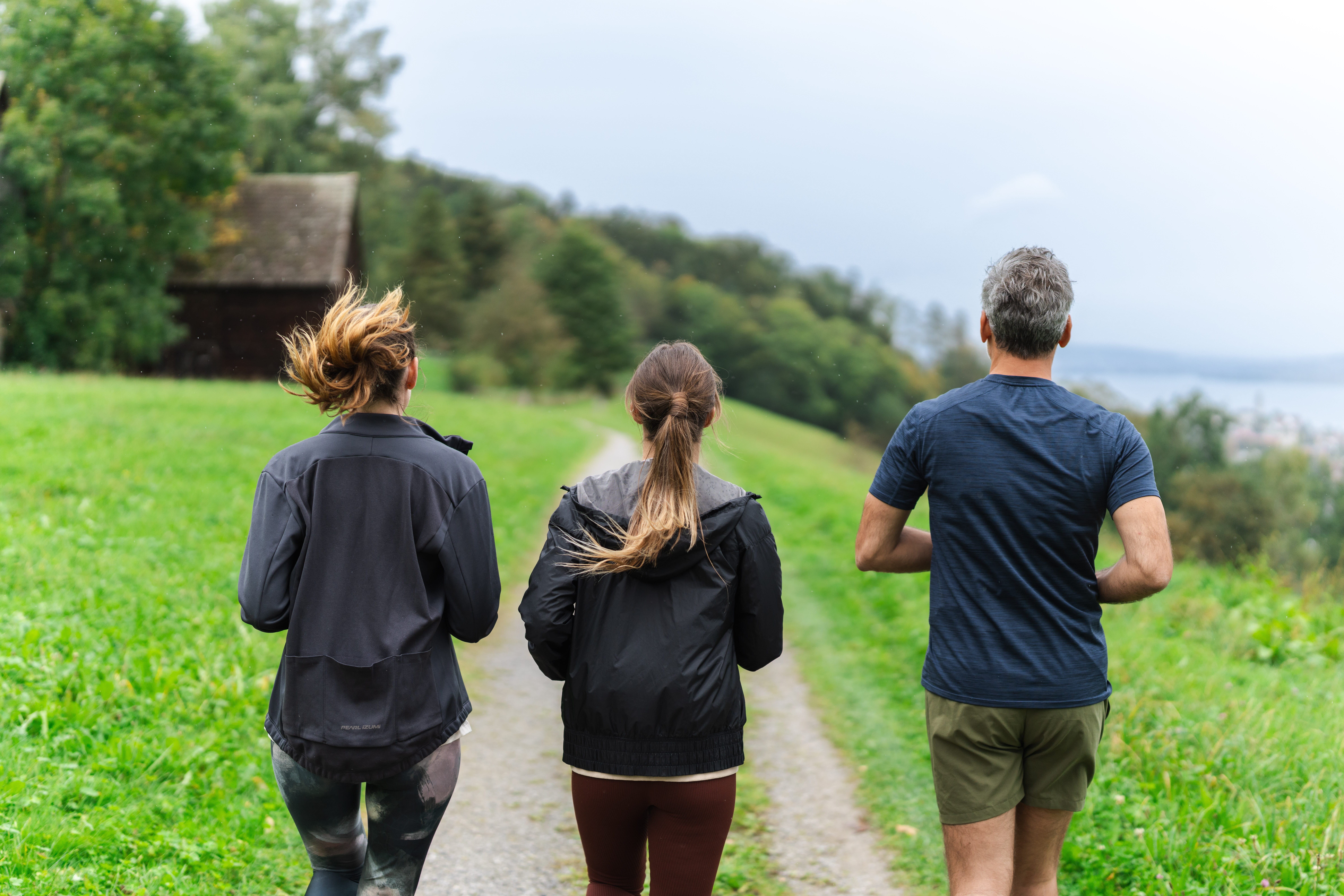 Drei Personen joggen auf einem Schotterweg durch eine grüne Landschaft, umgeben von Bäumen und Hügeln. Im Hintergrund ist ein Holzgebäude zu sehen. Sie sind von hinten in Sportkleidung zu sehen und genießen gemeinsam die Bewegung im Freien.