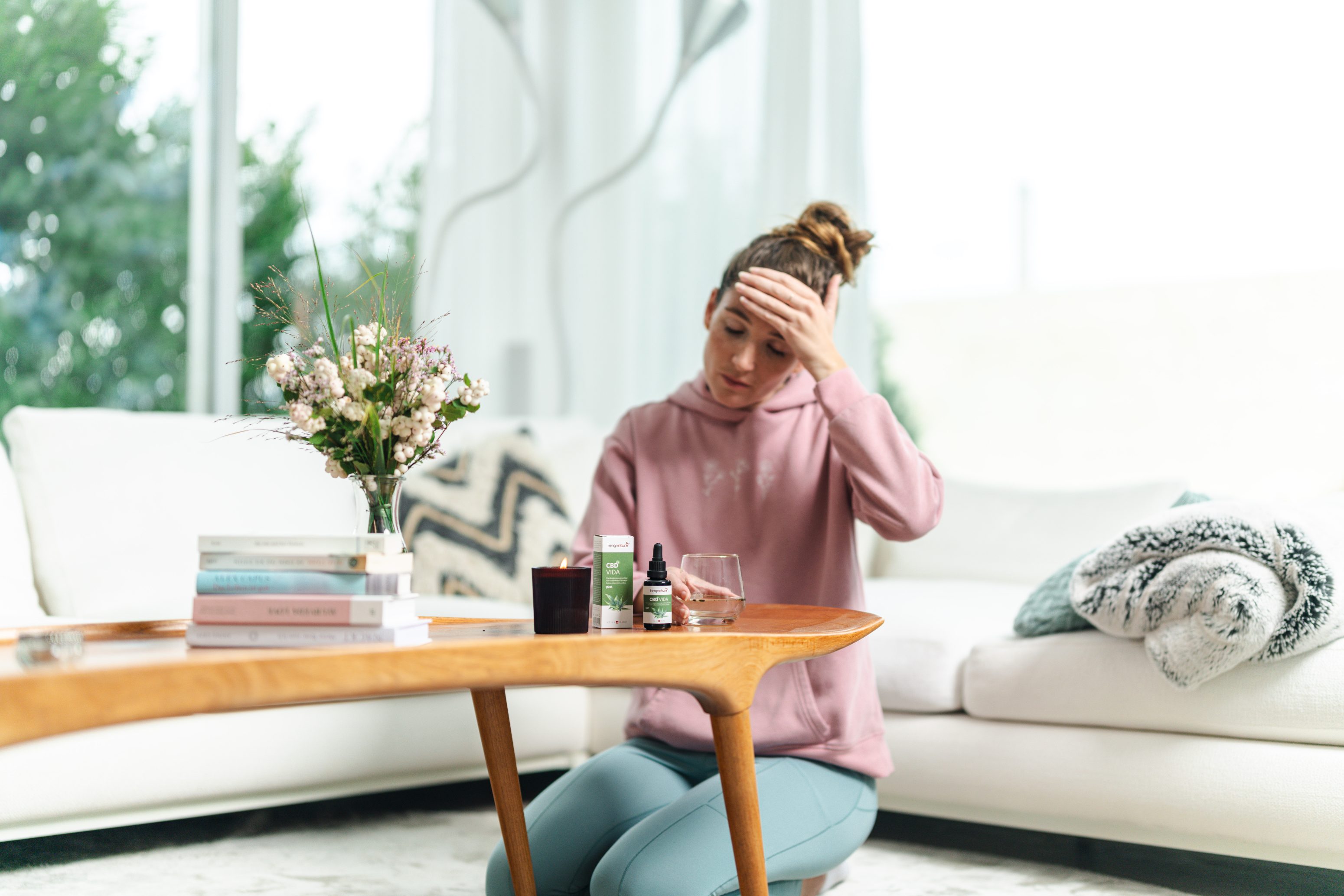 Eine Frau in einem rosa Kapuzenpullover und blauen Leggings sitzt auf dem Boden neben einem Couchtisch und hält sich die Stirn, als hätte sie Kopfschmerzen. Auf dem Tisch stehen Blumen, Bücher, eine Kerze, Nahrungsergänzungsmittel und ein Glas Wasser.