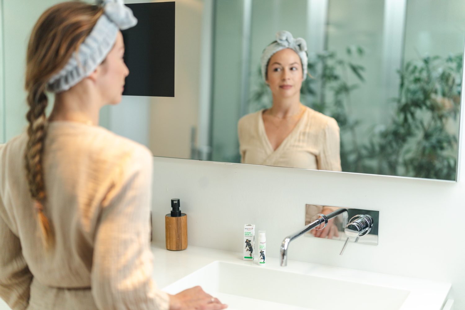 Eine Frau mit langen Haaren sitzt an einem Holztisch und liest ein Buch. Auf dem Tisch stehen eine Blumenvase, Glasbecher und dekorative Kerzenhalter. Durch die großen Fenster hinter ihr fällt sanft Sonnenlicht herein und schafft eine friedliche Atmosphäre.