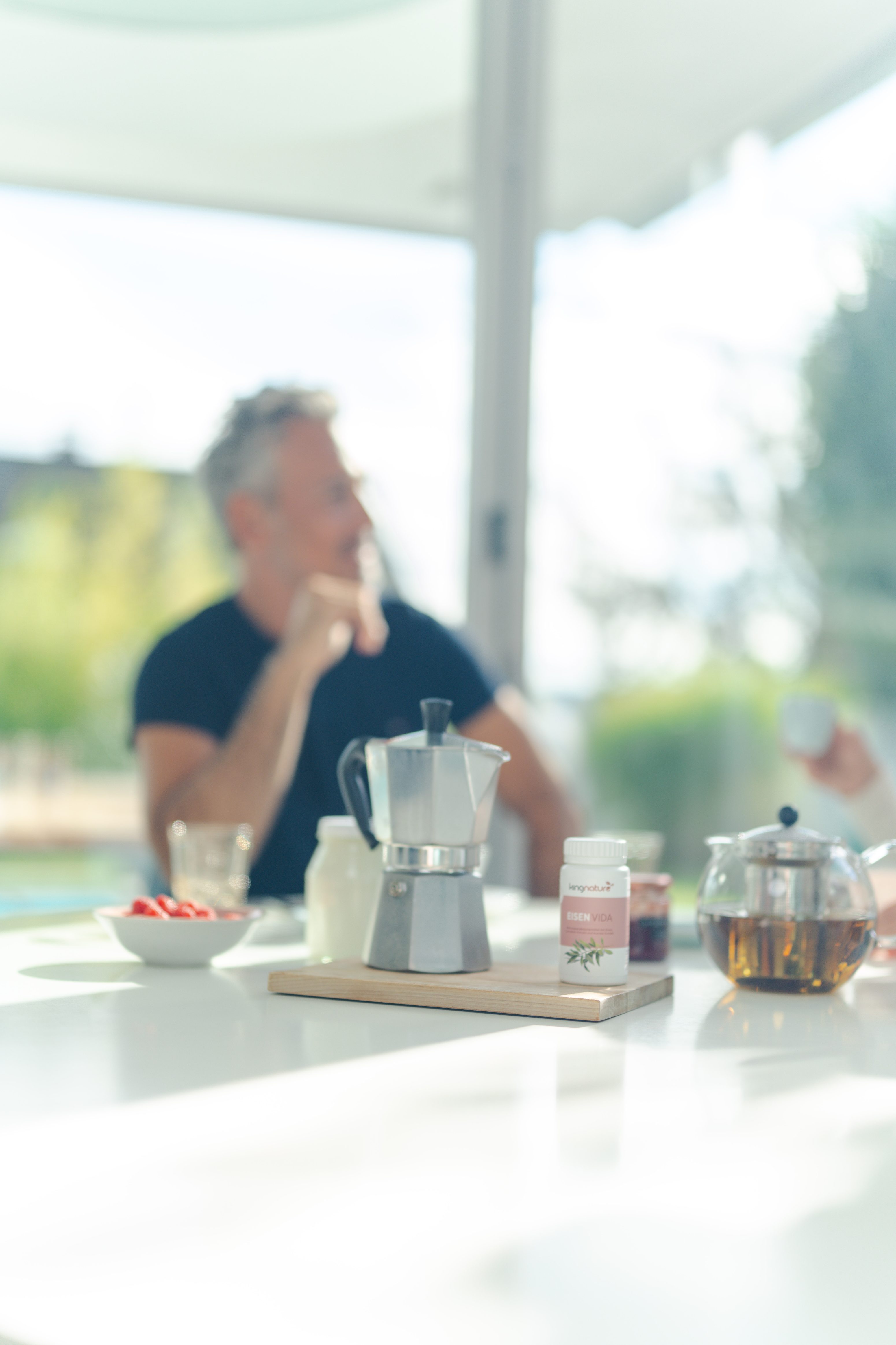 Ein verschwommener Mann sitzt an einem hellen Tisch. Im Vordergrund stehen eine Mokkakanne, eine Teekanne aus Glas, ein Glas und frische Erdbeeren. Sonnenlicht fällt durch große Fenster herein.