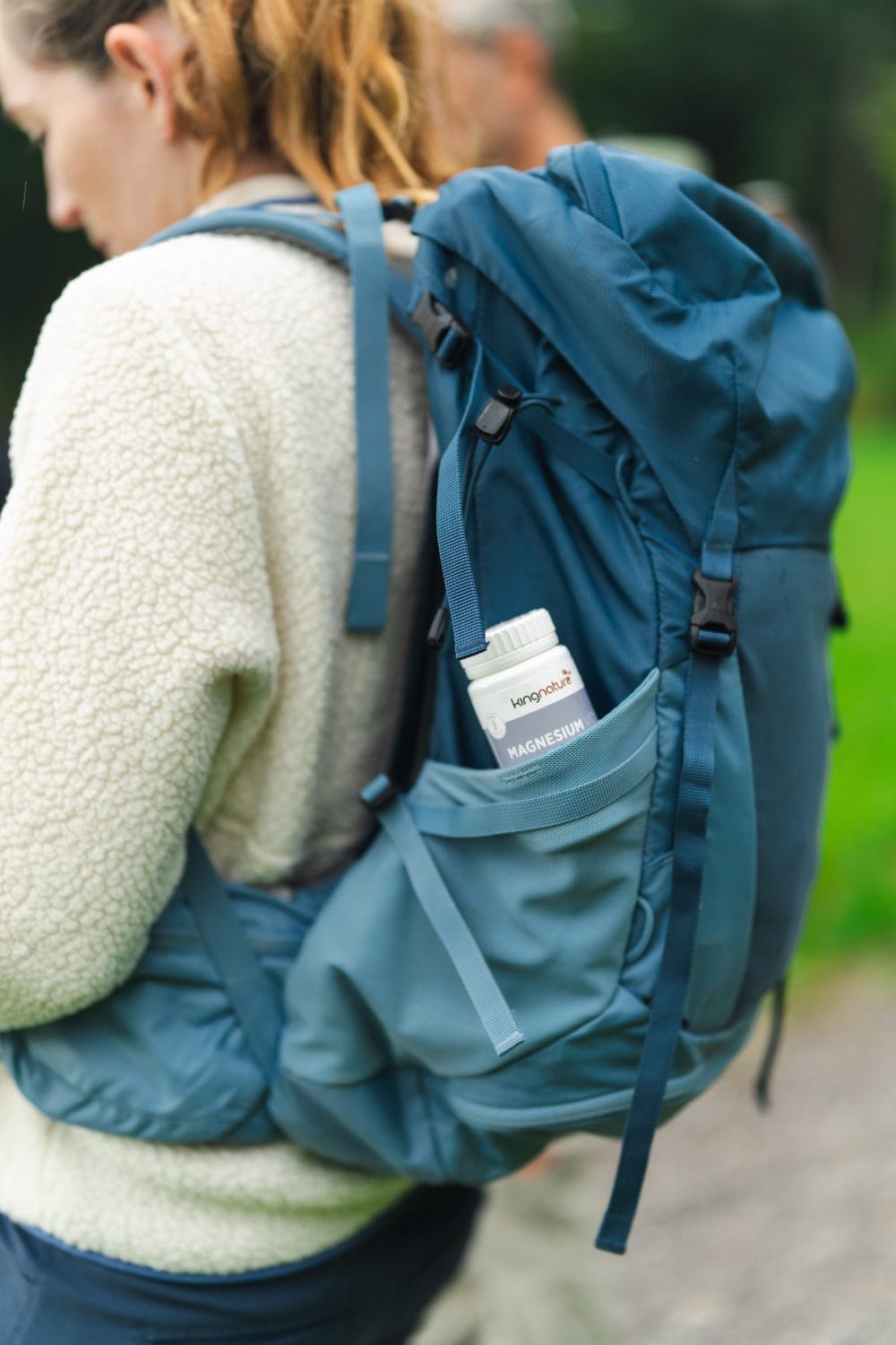 Eine Person trägt einen blauen Wanderrucksack, in dessen Seitentasche sich eine weiße Flasche mit der Aufschrift „Magnesium Kapseln“ befindet. Bekleidet mit einer cremefarbenen Fleecejacke steht die Person im Freien, umgeben von Grün.