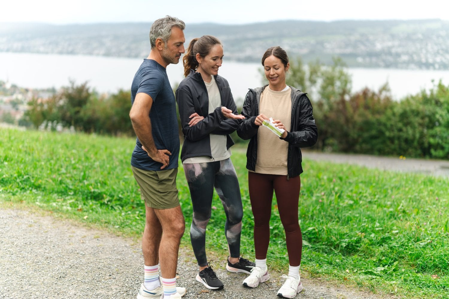 Drei Personen in Sportkleidung unterhalten sich draußen auf einem Weg an einem grünen Rasen und einem See. Eine Frau hält eine Wasserflasche in der Hand, während die anderen sie beobachten. Alle lächeln und wirken entspannt nach einem gemeinsamen, energiegeladenen Training.